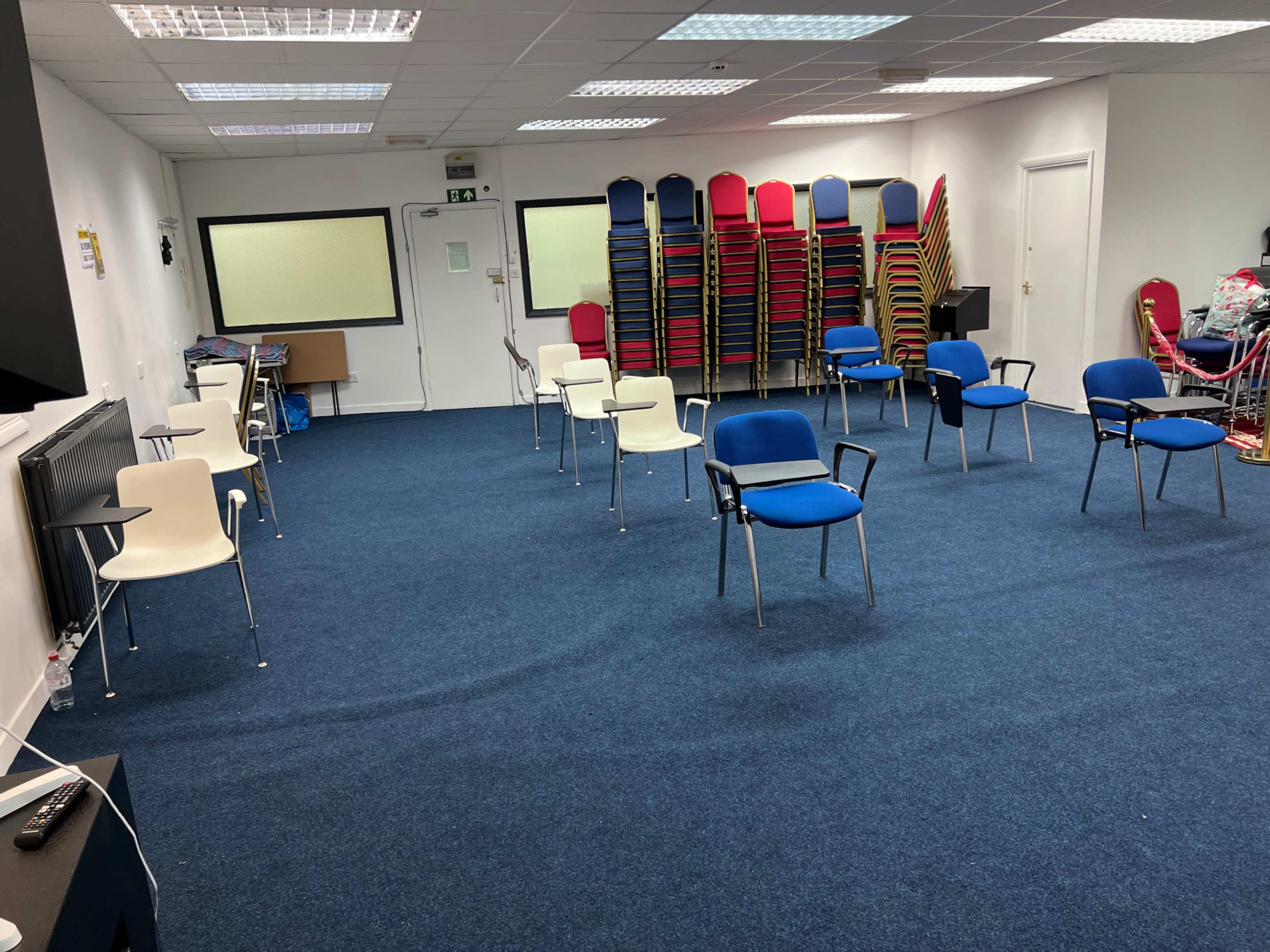 The image shows a room with blue carpet, several empty chairs arranged in pairs, and a stack of colorful chairs against the wall.