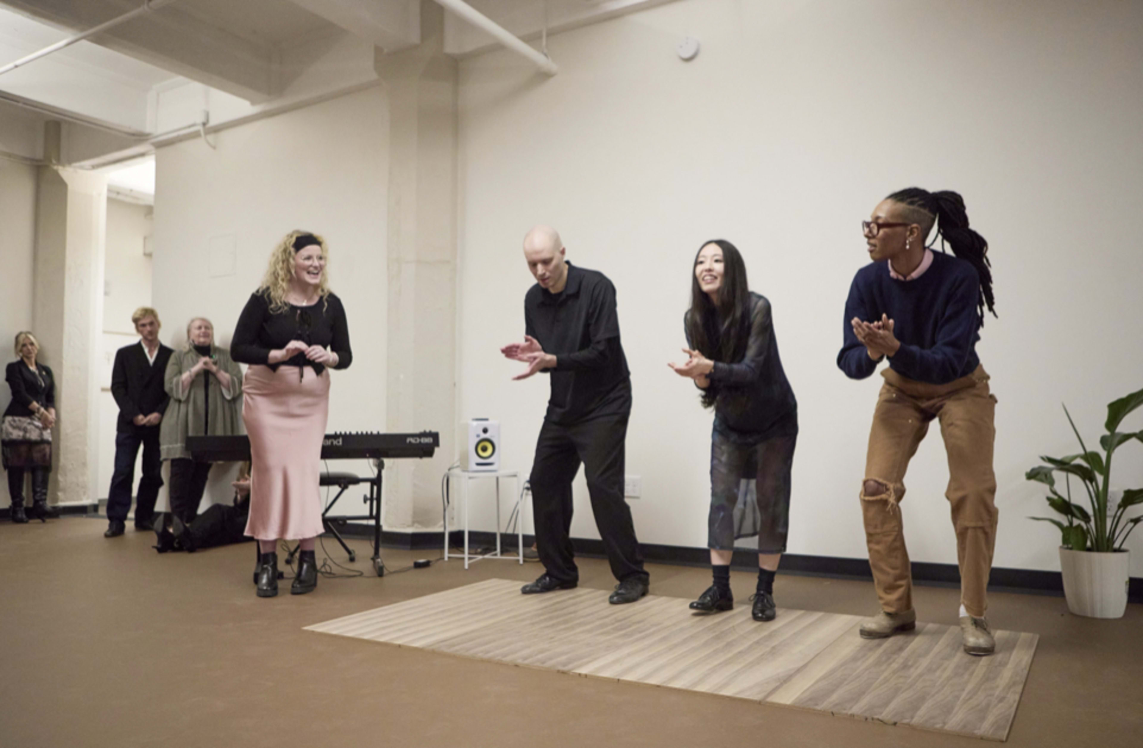 A group of four performers stands on a wooden platform, clapping, while an audience observes from the background.