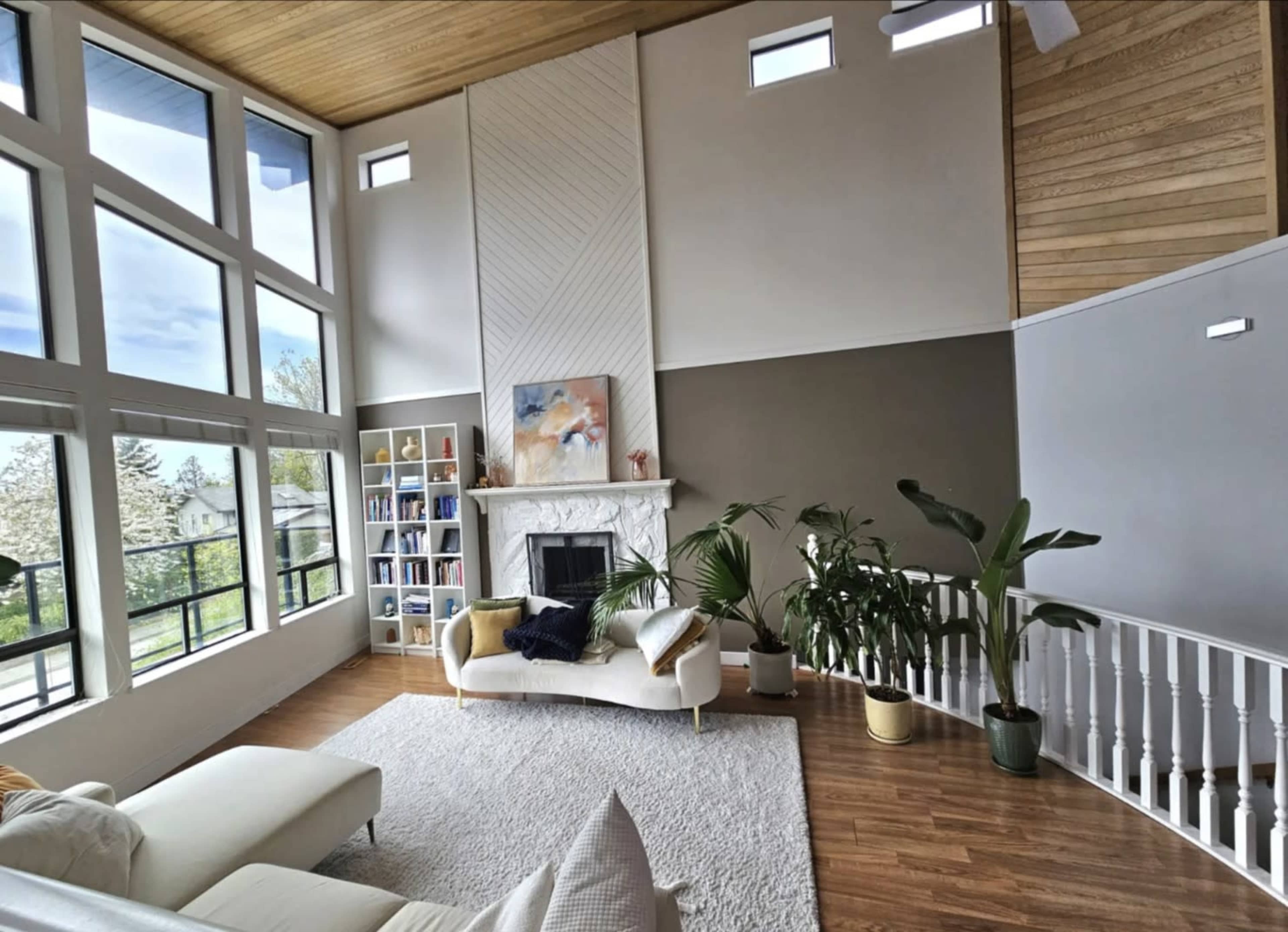 The image shows a spacious living area with large windows, a fireplace, a white sofa, a bookshelf, and indoor plants, all under a wooden ceiling.
