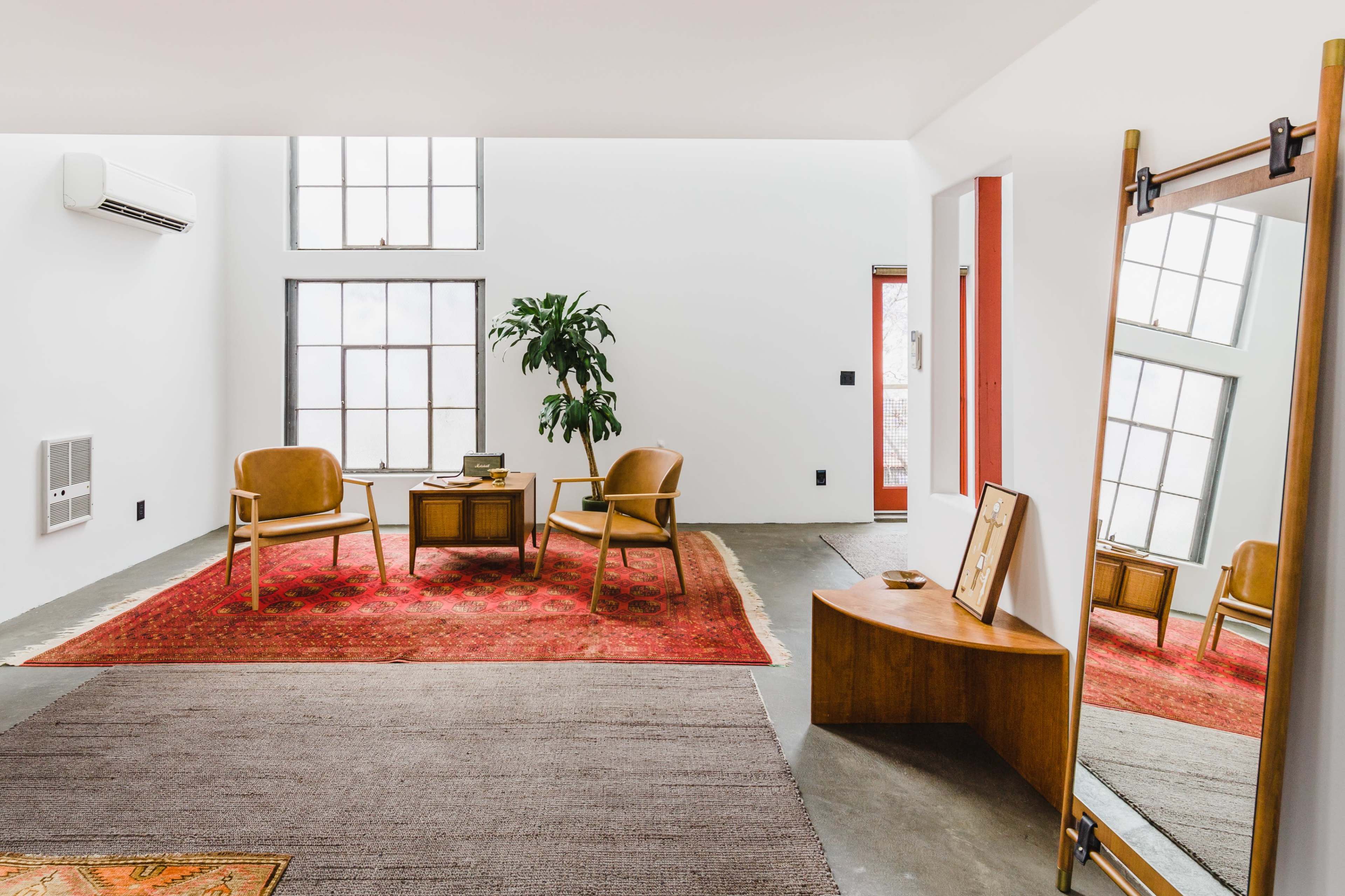 A modern living room features two chairs, a coffee table, a large mirror, and a plant, all set against a backdrop of white walls and large windows.