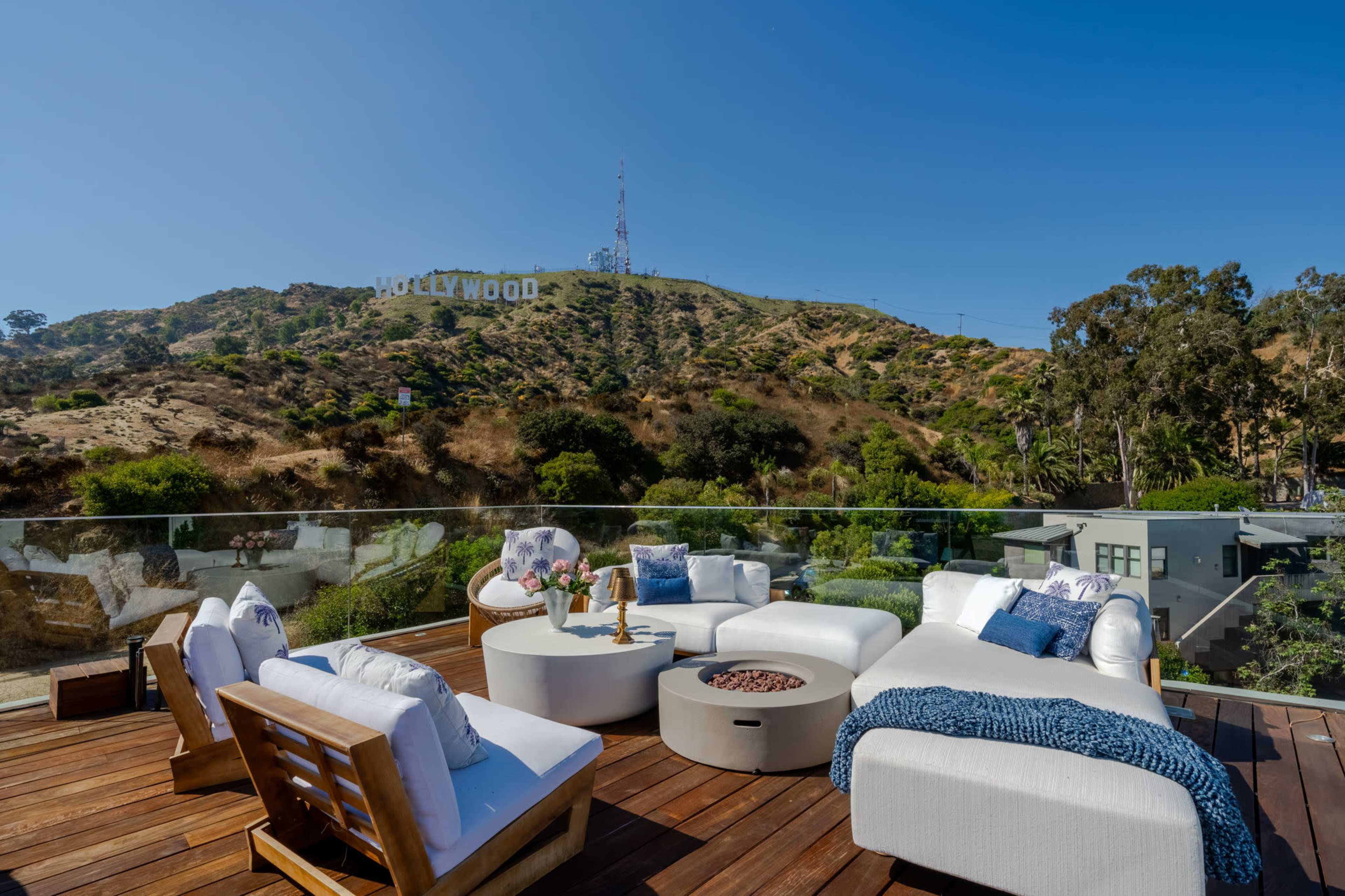 A rooftop patio features modern seating and a fire pit with a view of the Hollywood sign on the hillside.
