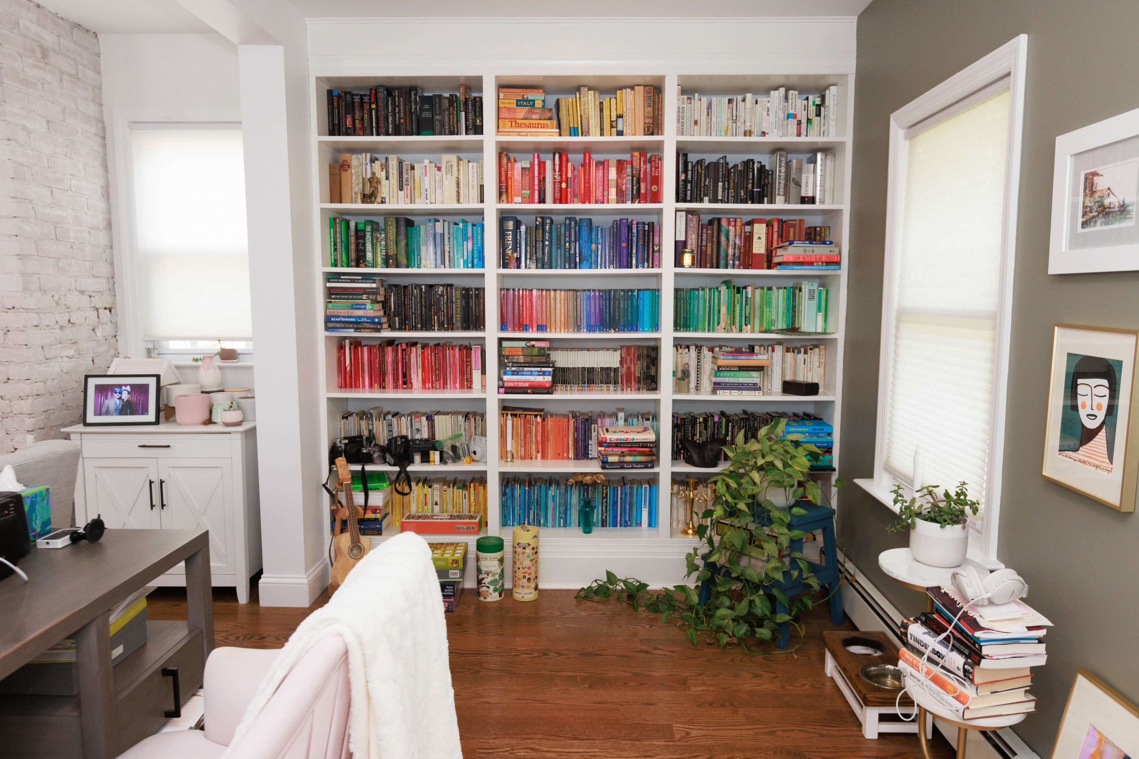 A large bookshelf filled with books organized by color stands against a wall in a well-lit room, accompanied by a small plant and a desk in the foreground.