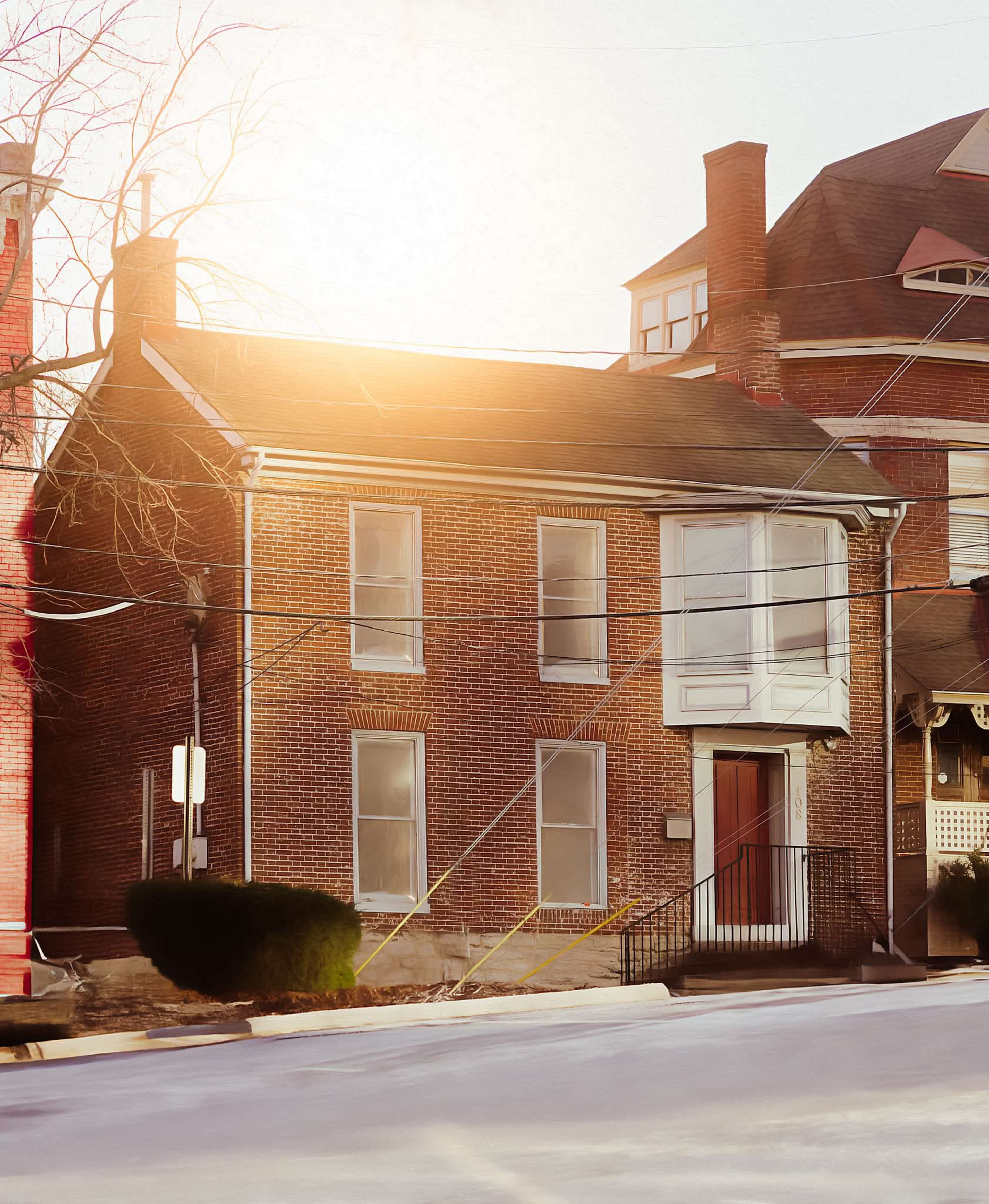 The image shows a brick house with white trim located at the corner of a street, with power lines visible in the foreground.