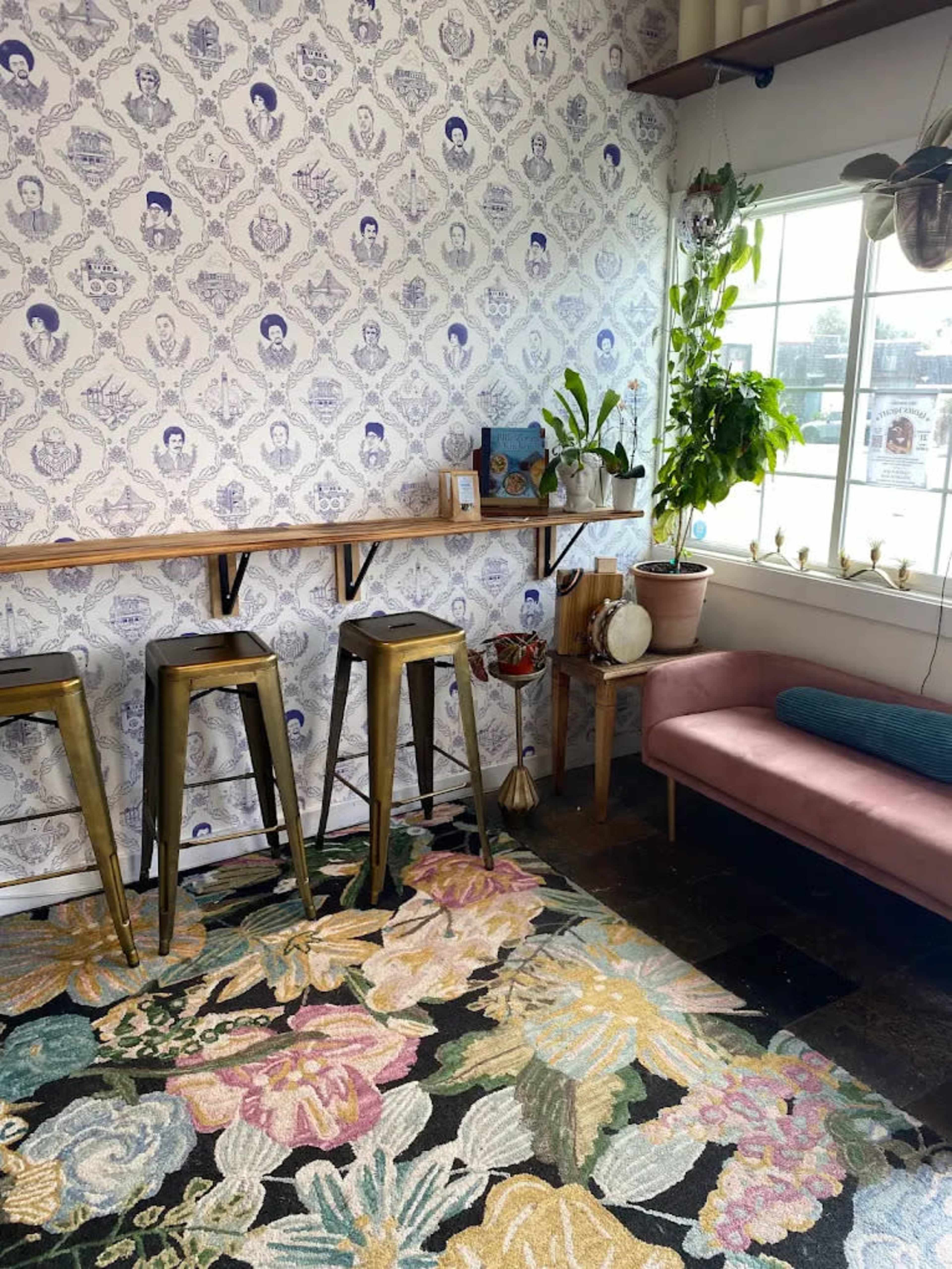 The image shows a brightly lit corner of a room featuring a patterned wallpaper, a wooden bar with metallic stools, a pink couch, a decorative rug with floral patterns, and several indoor plants.