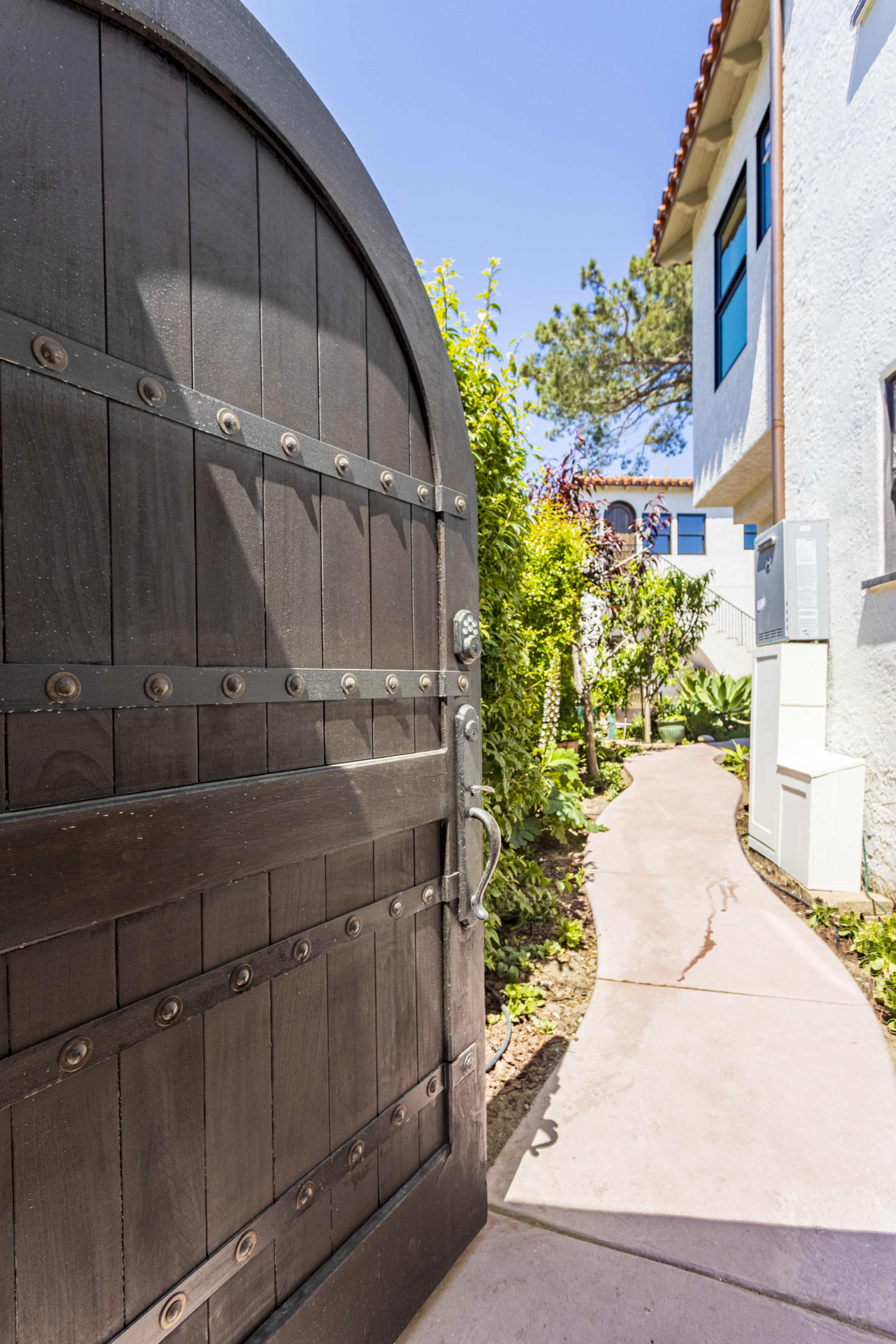 A black wooden gate stands open, revealing a landscaped pathway leading between a white building and greenery.