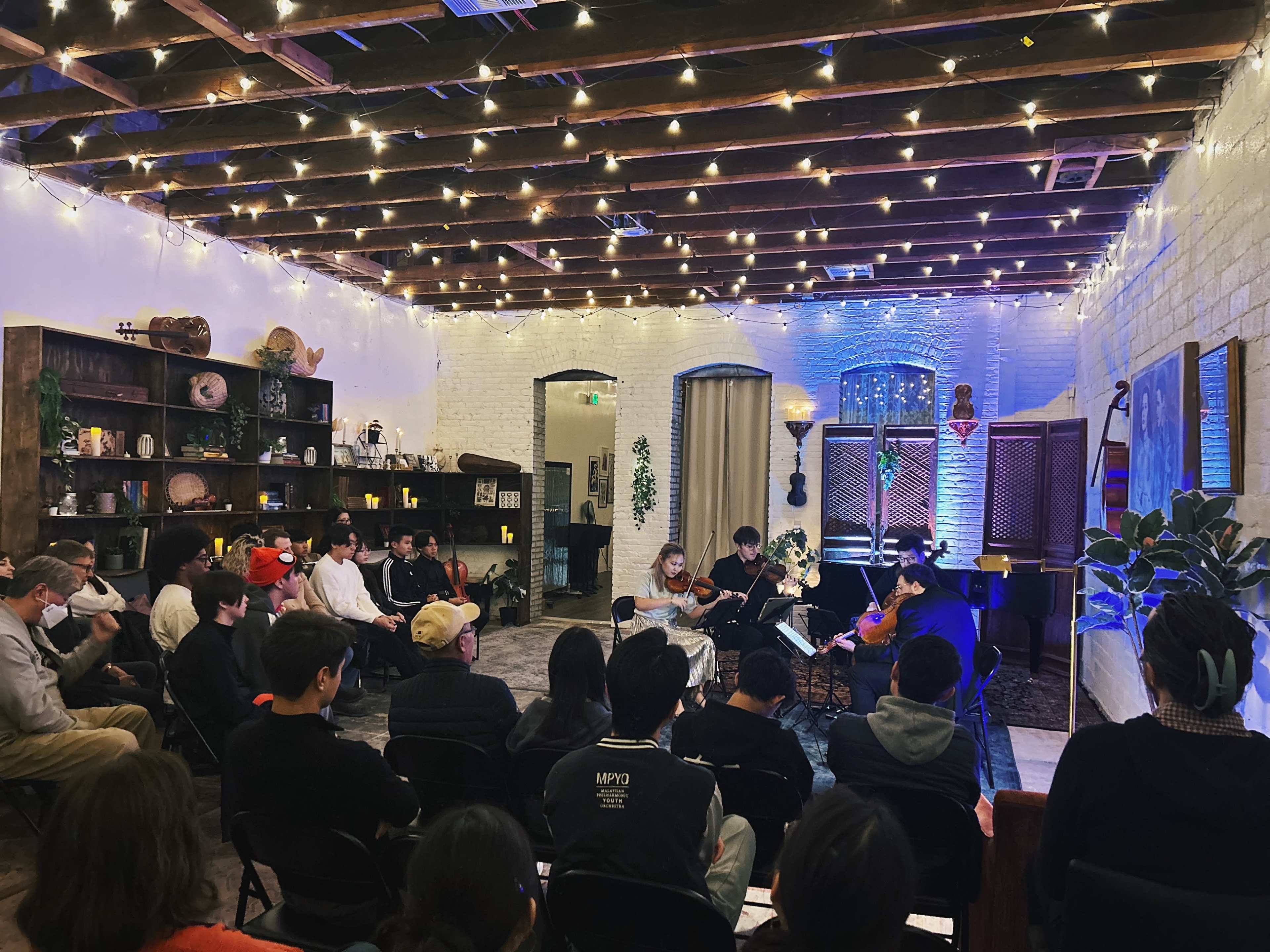 A group of people sits in a warmly lit room with string lights, listening to musicians performing in front of a decorative backdrop.