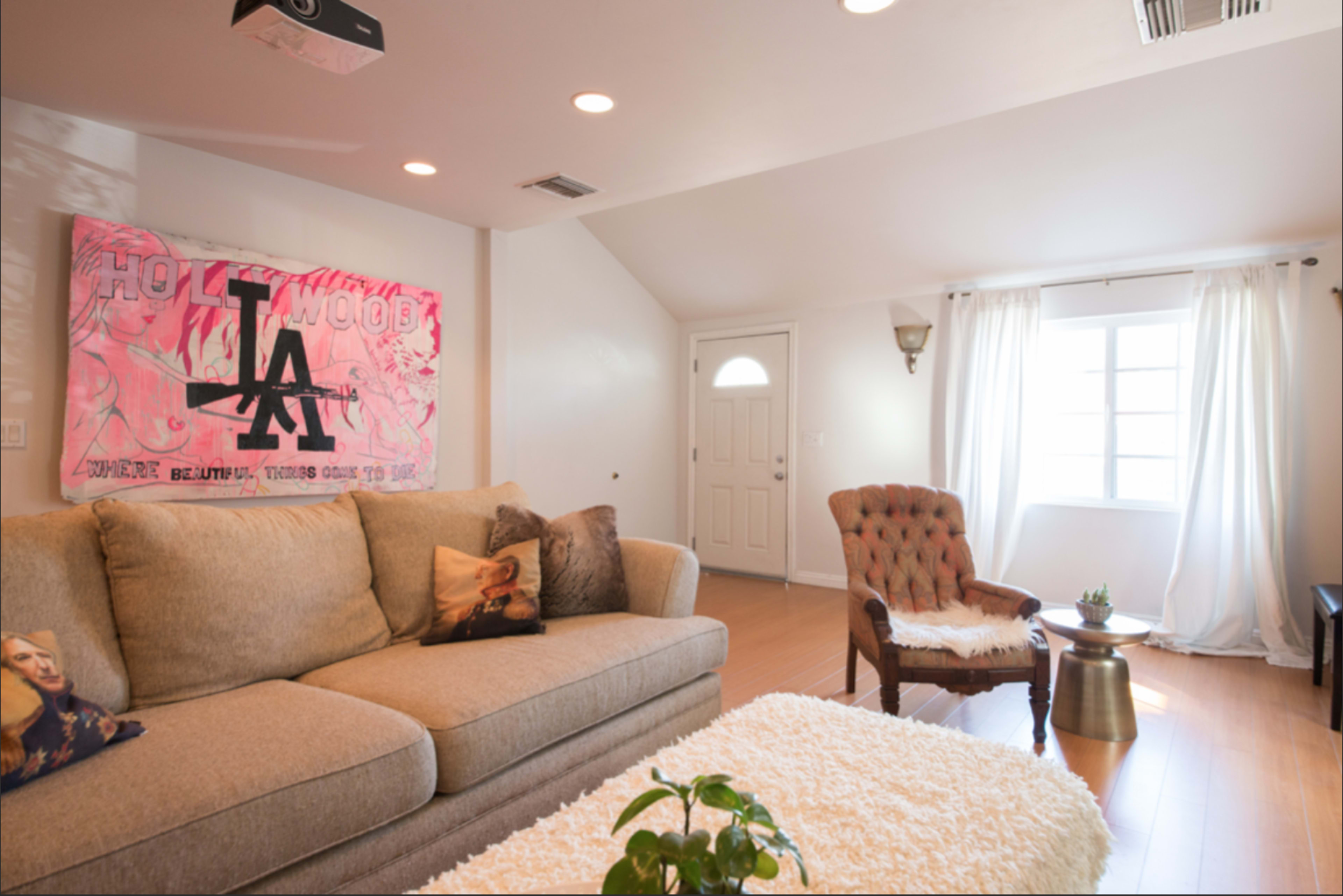 A living room features a beige couch with decorative pillows, a wooden armchair, a coffee table with a plant, and artwork on the wall, along with a window letting in natural light.