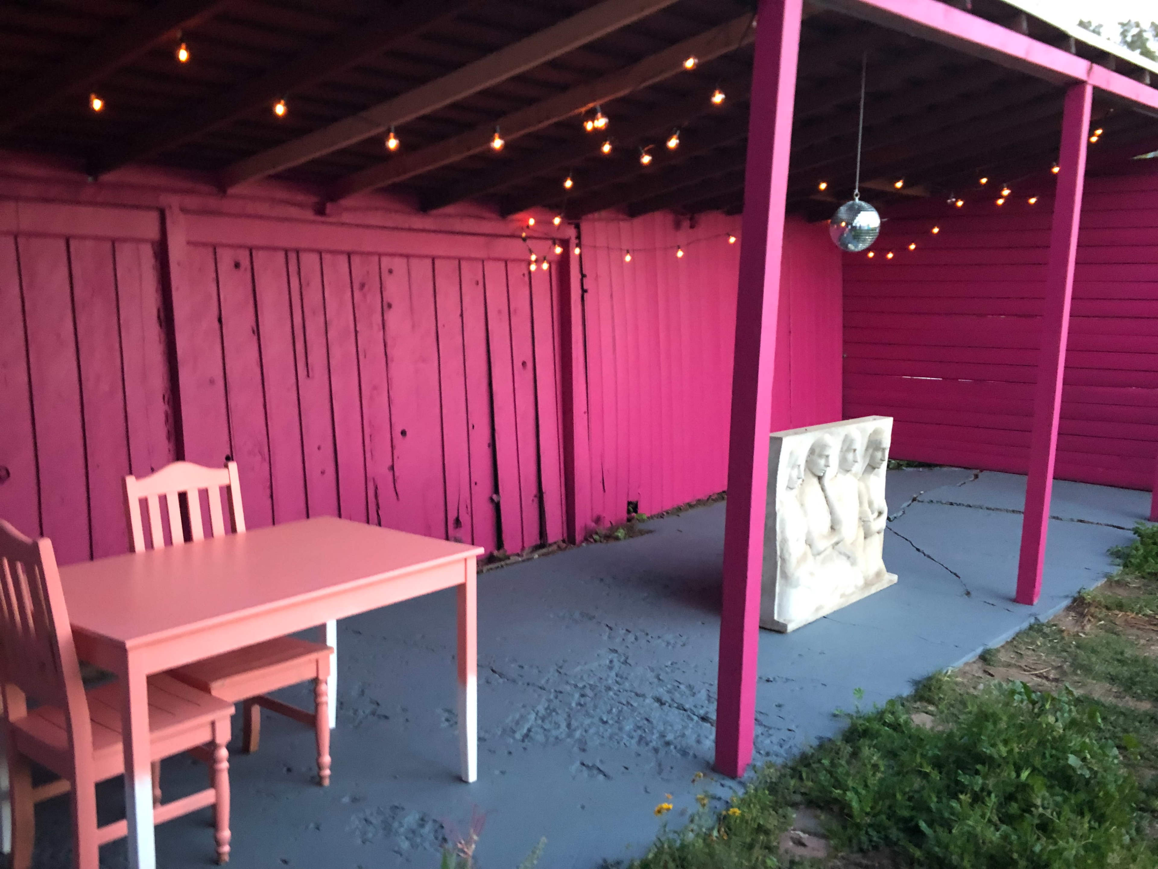 A small outdoor space featuring a pink wooden wall, a white table with matching chairs, and a sculpture of three figures, all illuminated by string lights overhead.