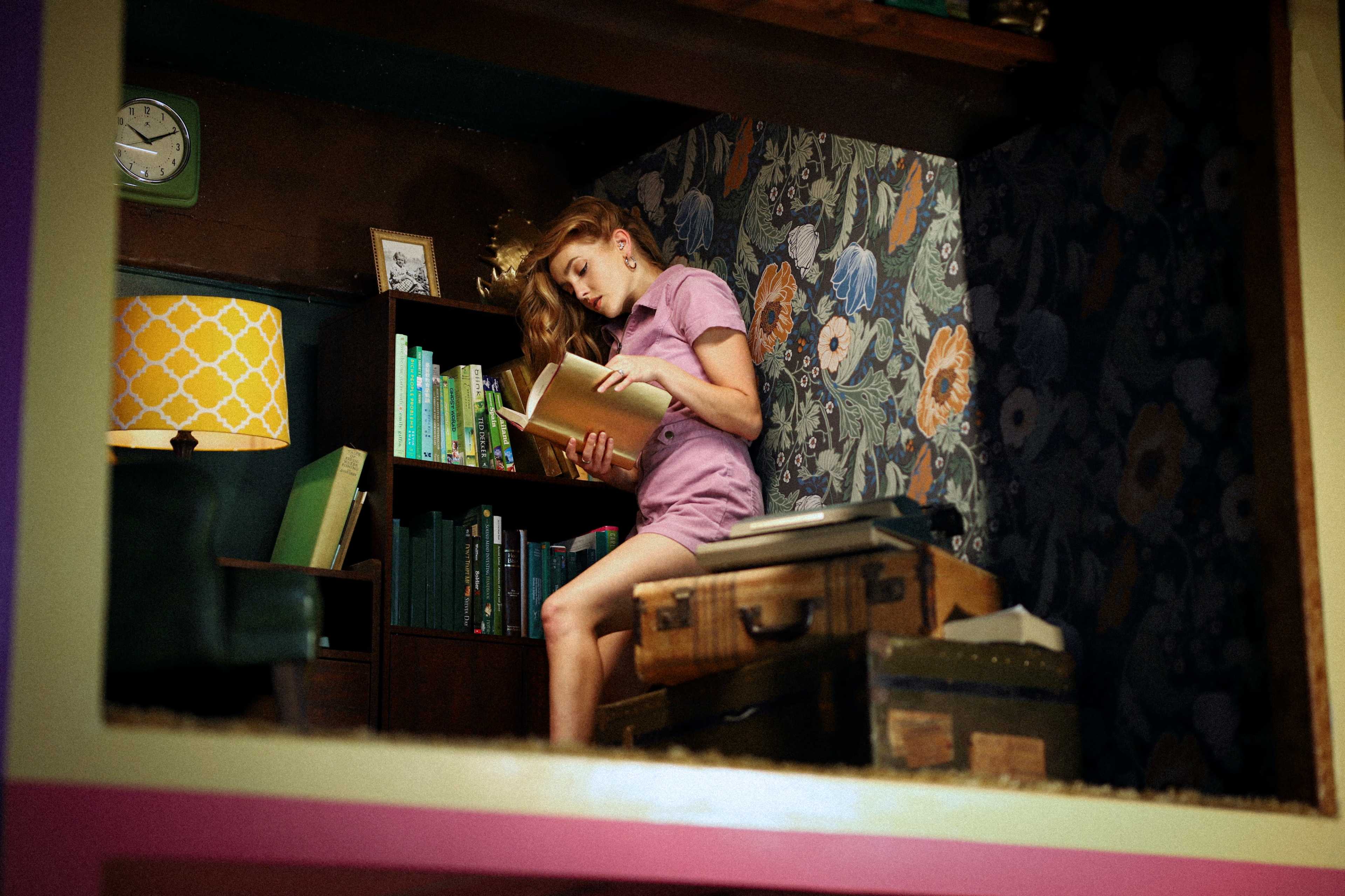 A young woman in a pink outfit sits on a wooden box, reading a book in a cozy, book-filled room with floral wallpaper.