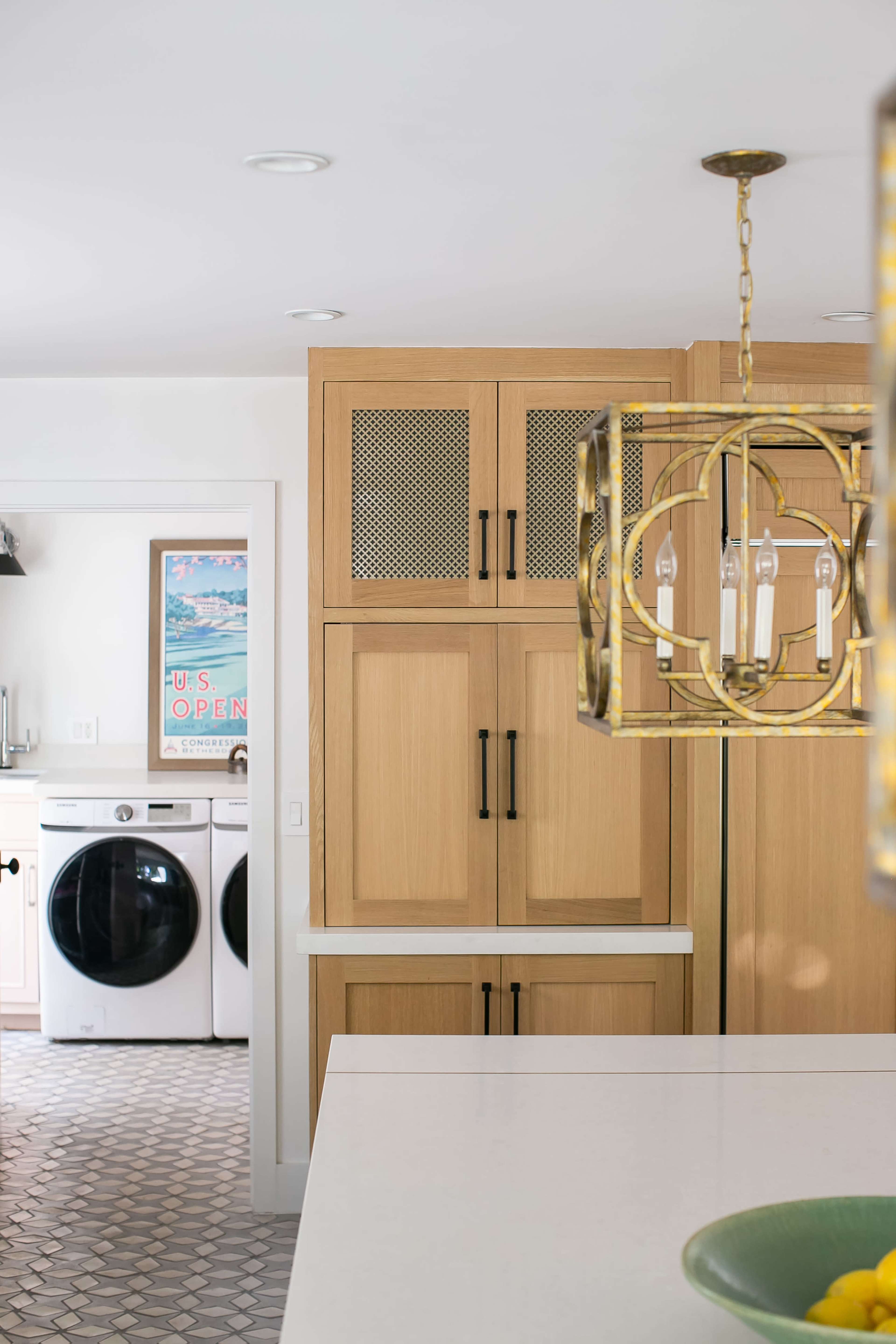 A modern kitchen features wooden cabinetry, a dining area with a chandelier, and a laundry room visible through an open doorway.