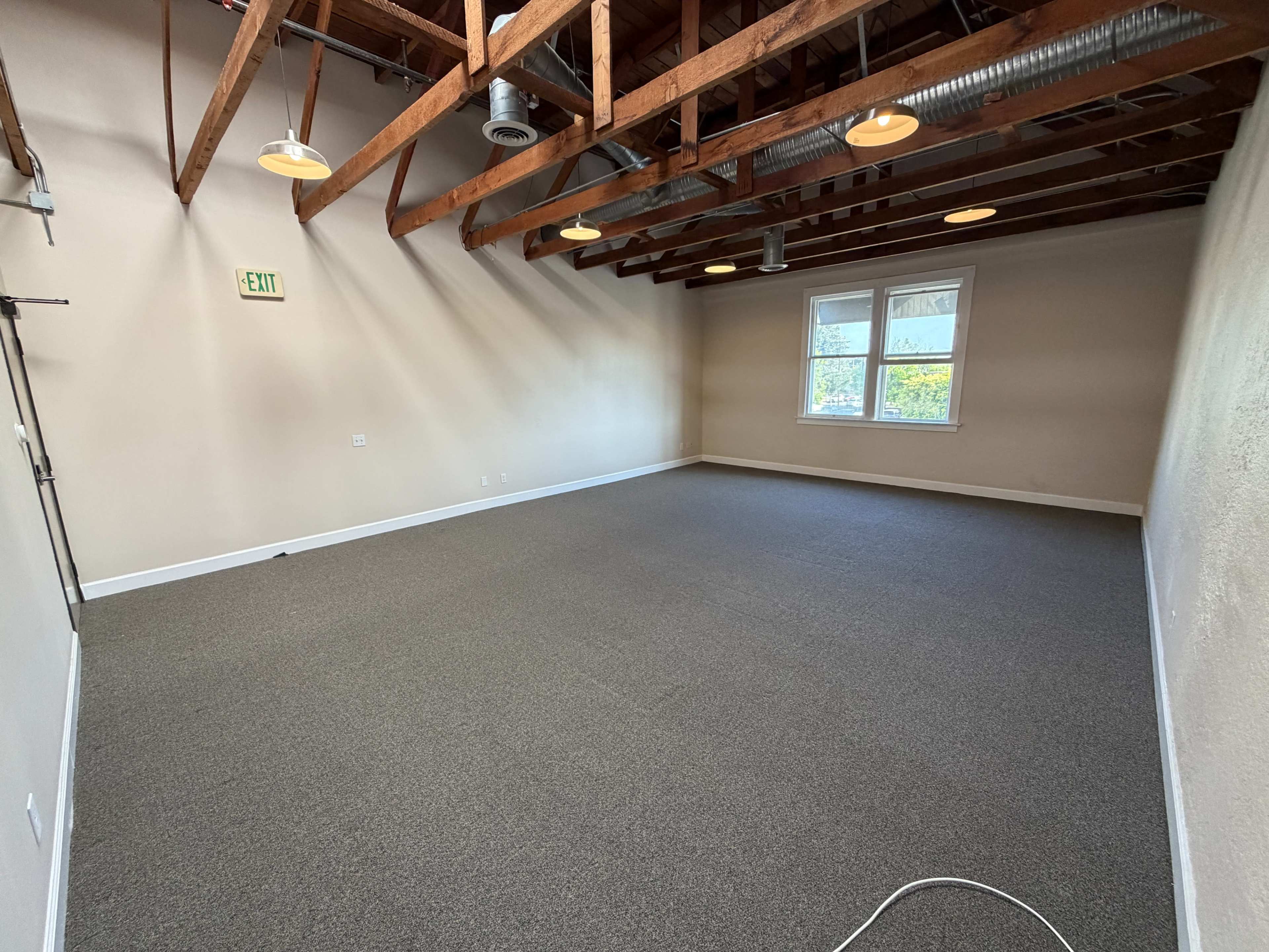The image shows a spacious, empty room with exposed wooden beams and gray carpet flooring.