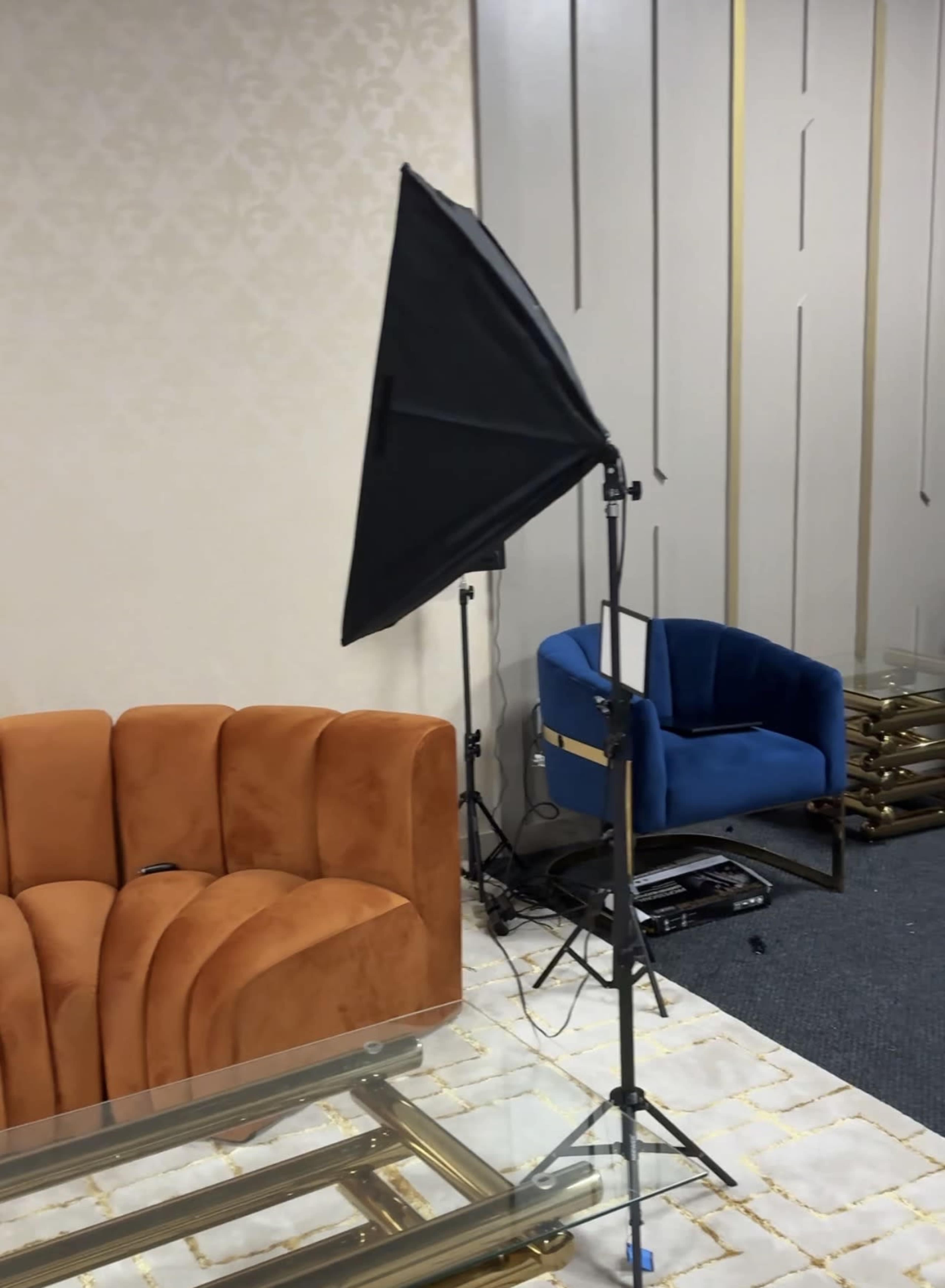 The image shows an orange velvet couch, a blue armchair, and a lighting setup on a stand in a room with light-colored walls and a patterned floor.