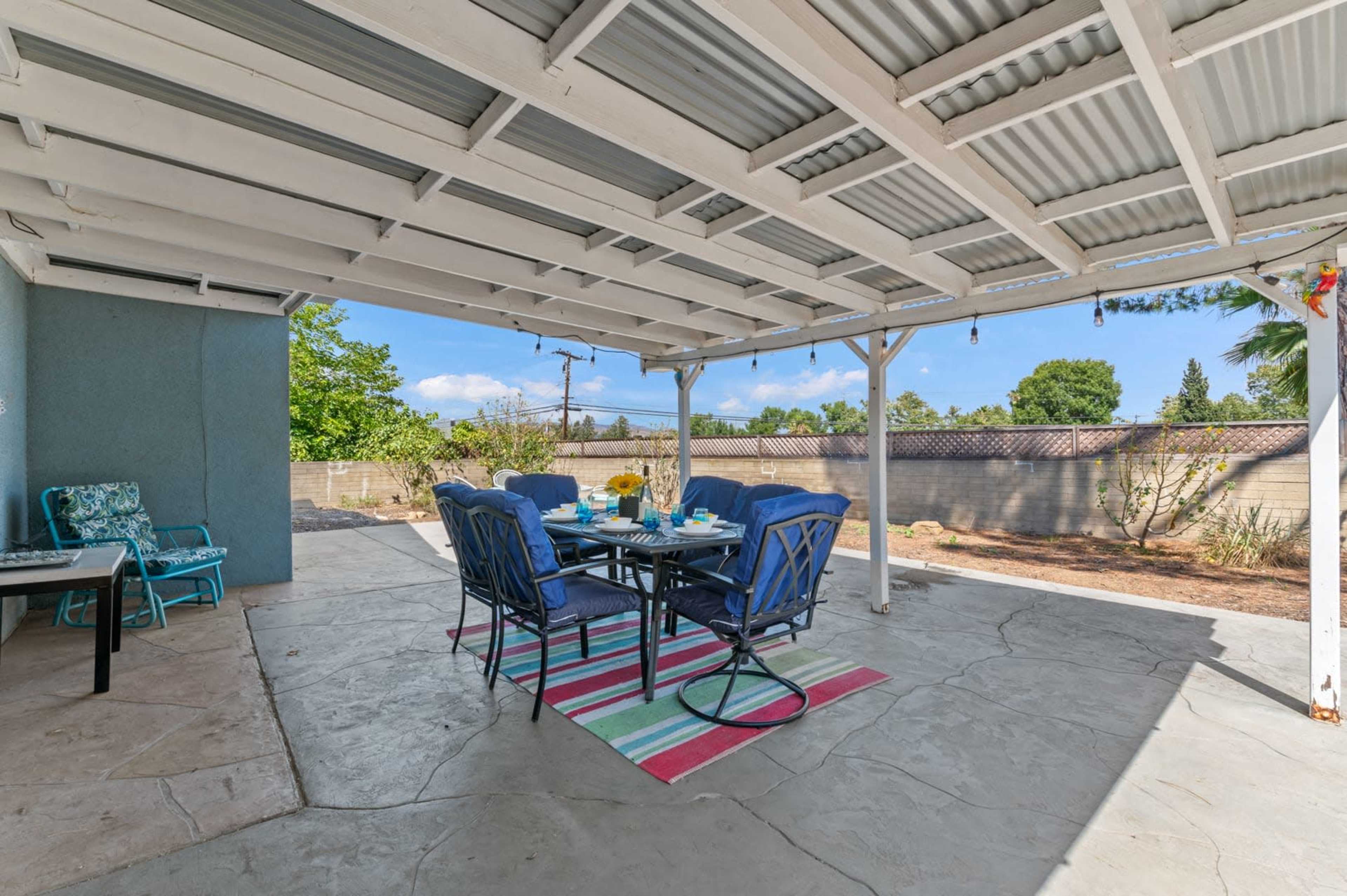 A covered patio with a dining table and chairs is set up on a concrete surface, surrounded by a landscaped yard.