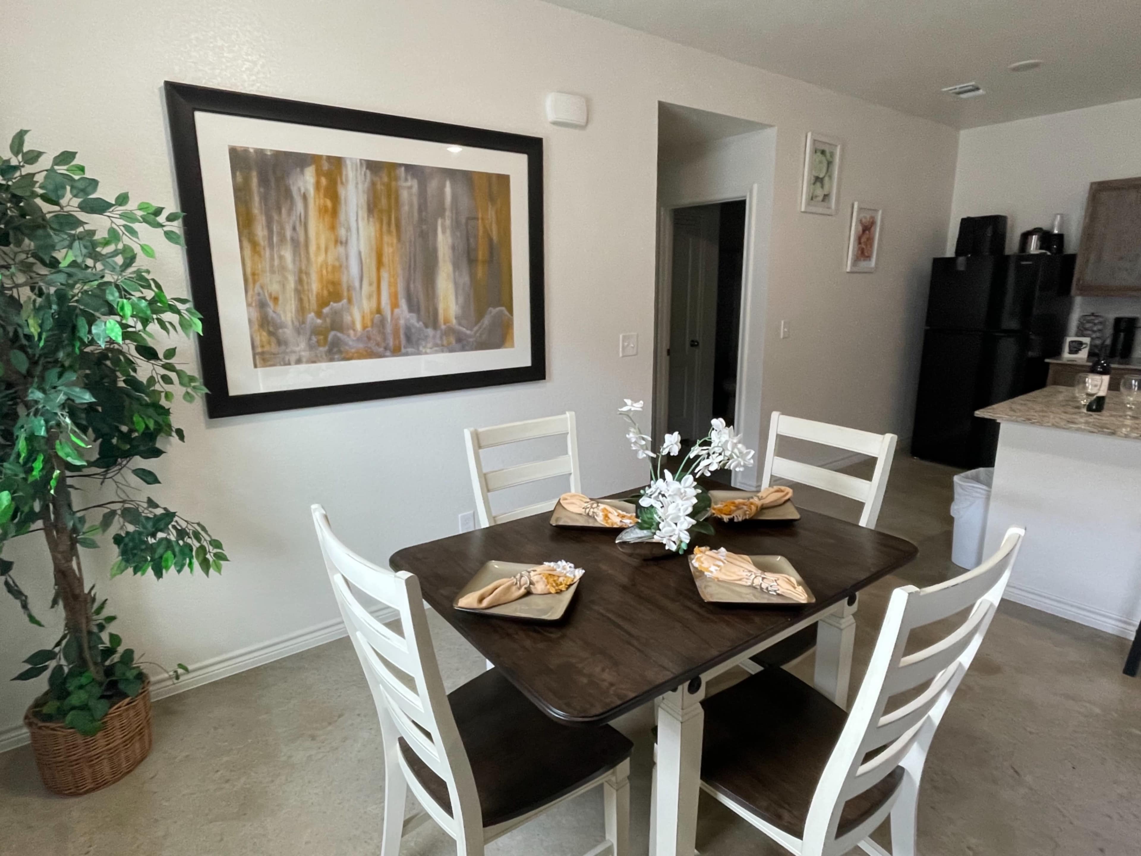 A dining table set with decorative napkins and flowers is positioned near a framed artwork in a kitchen area.