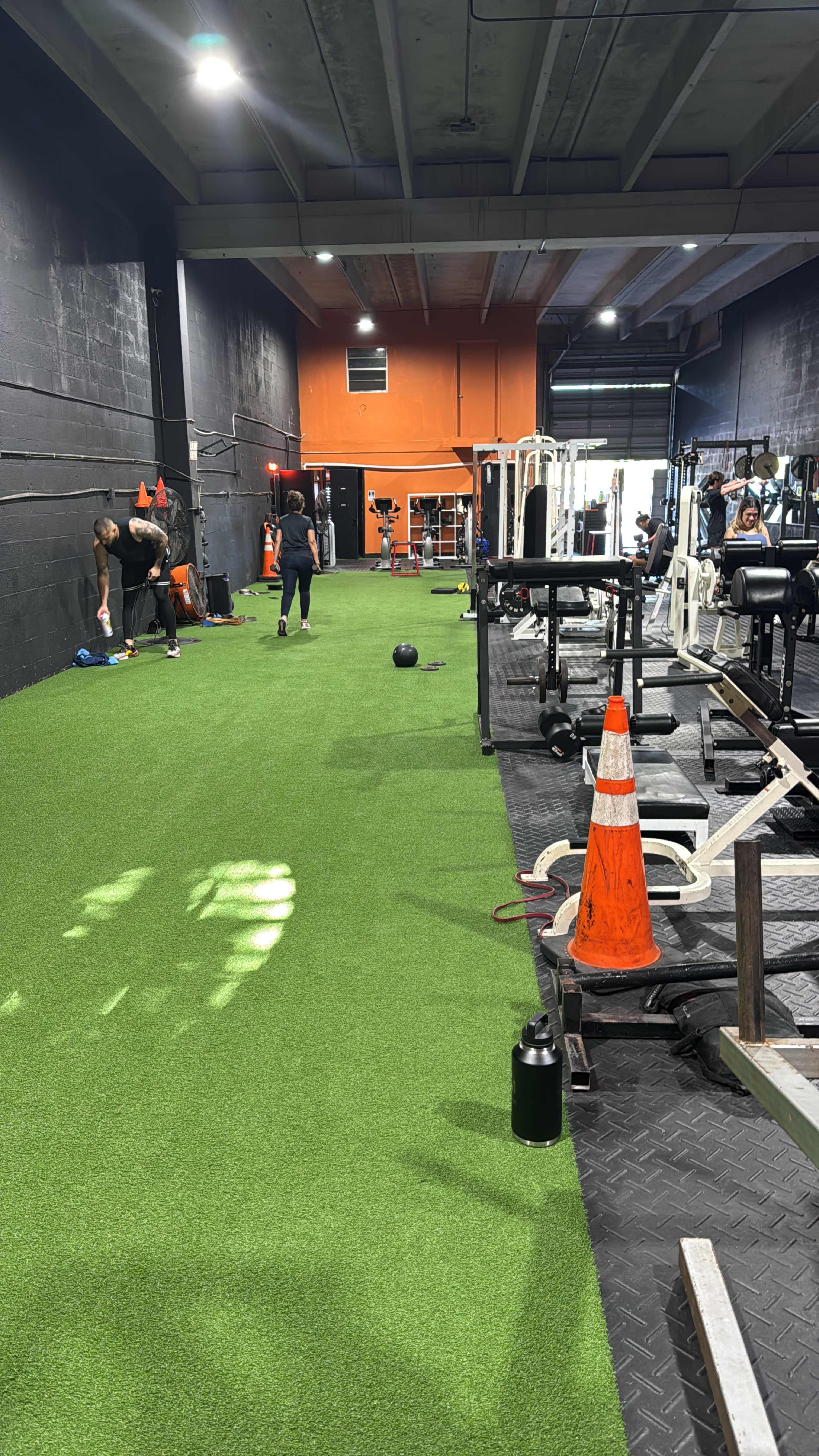 The image shows a gym interior with artificial turf flooring, workout equipment along the sides, and individuals engaged in exercise activities.