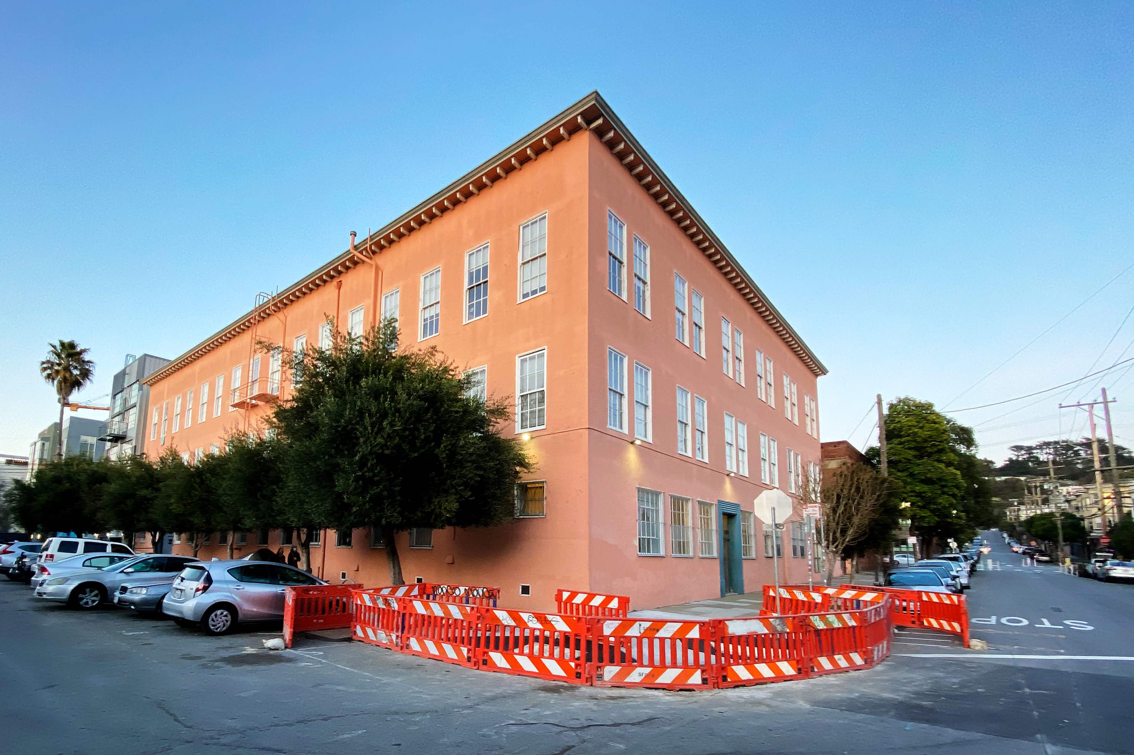 A pink building with large windows is located at the corner of a street, surrounded by orange construction barriers and several parked cars.