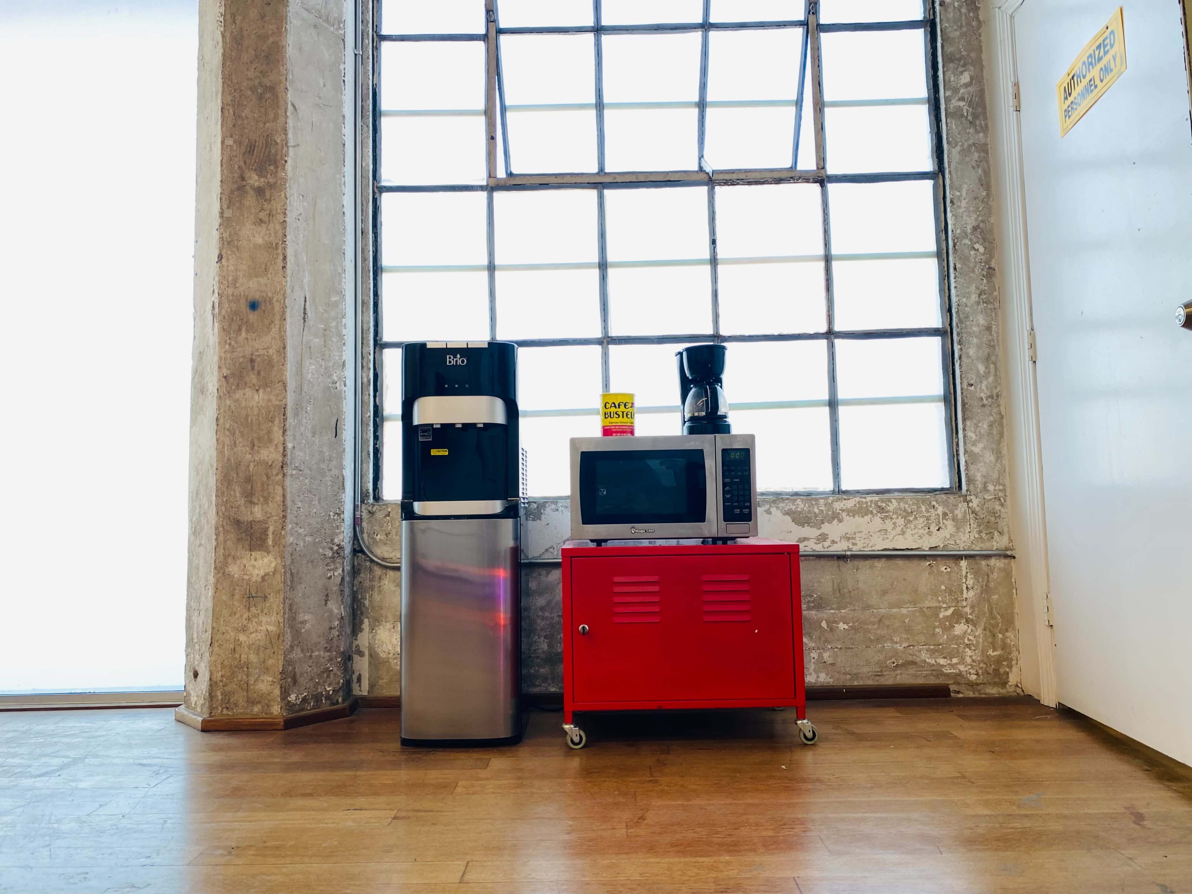 A stainless steel refrigerator and a microwave sit beside a red cart, all placed against a large industrial window.