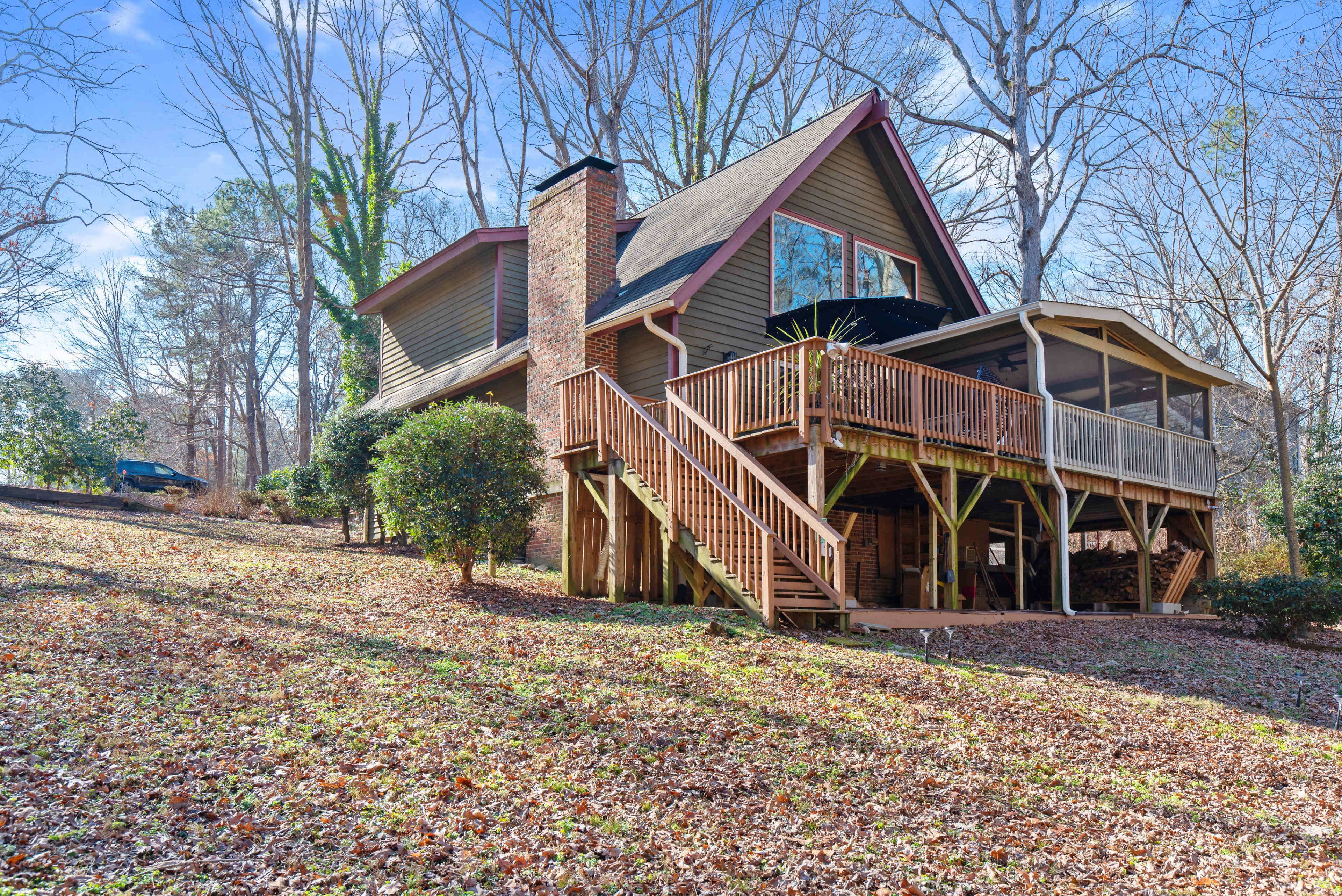 A two-story house with a large deck situated on a sloped yard covered in fallen leaves, surrounded by trees.