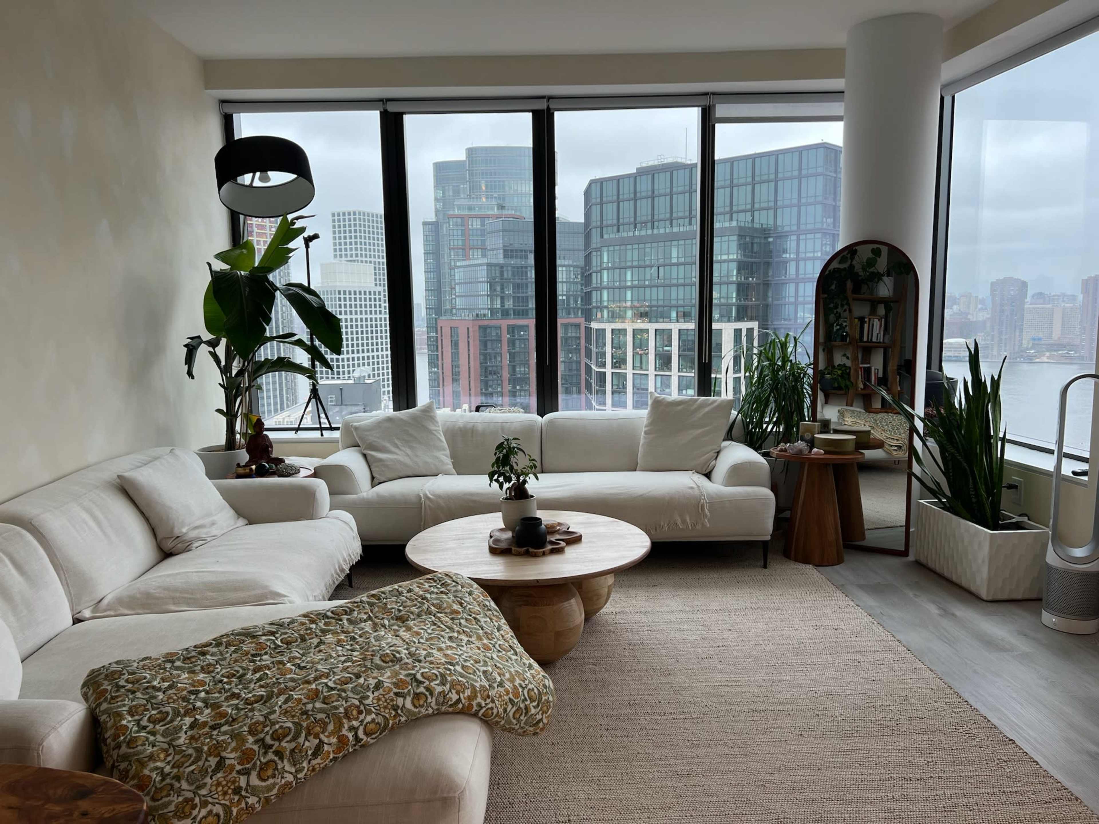 A modern living room features a light-colored sectional sofa, a round wooden coffee table, and large windows showcasing a city skyline.