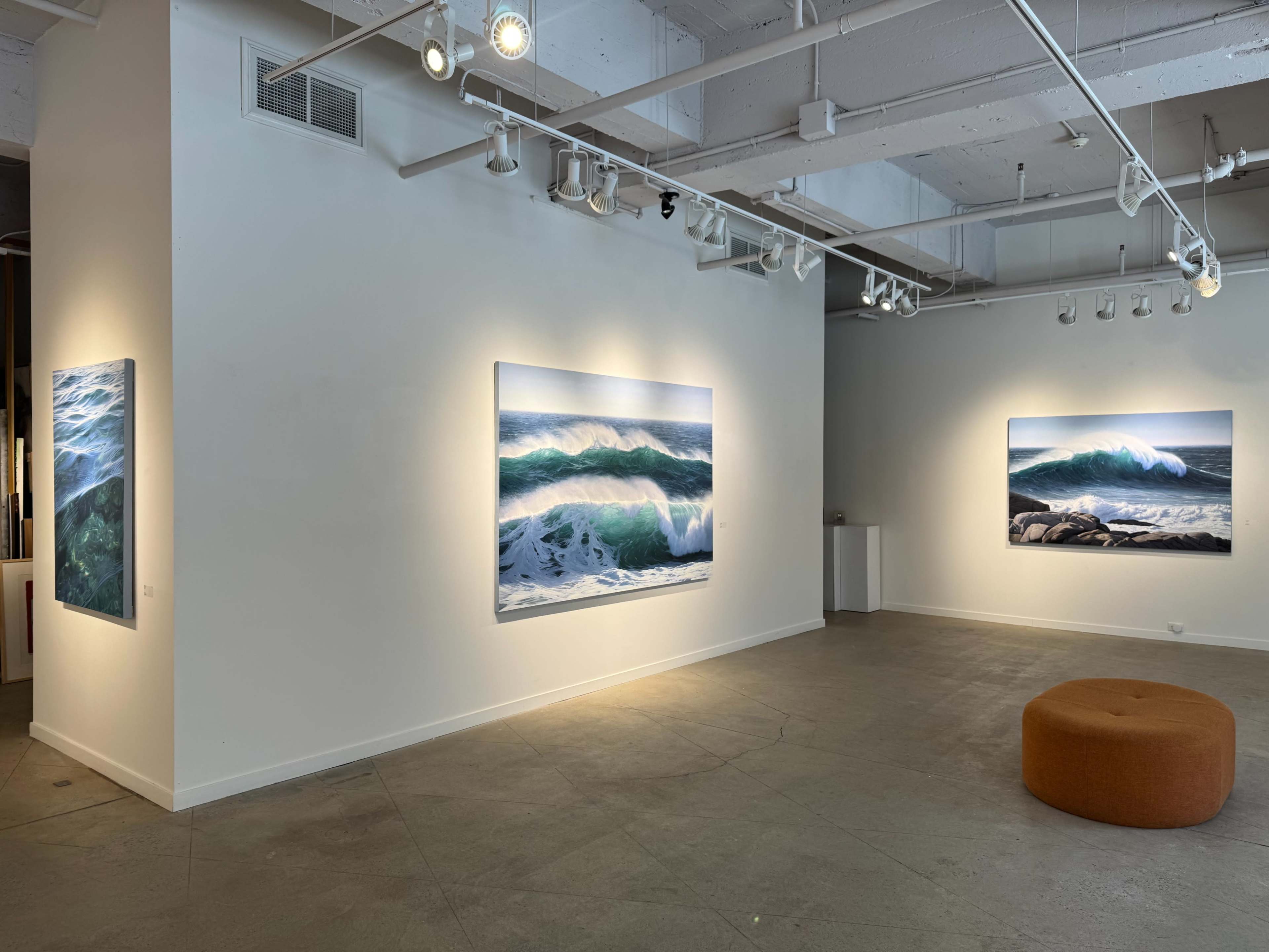 The image shows an art gallery displaying three large ocean wave photographs on the walls, with a circular orange seating area in the center.