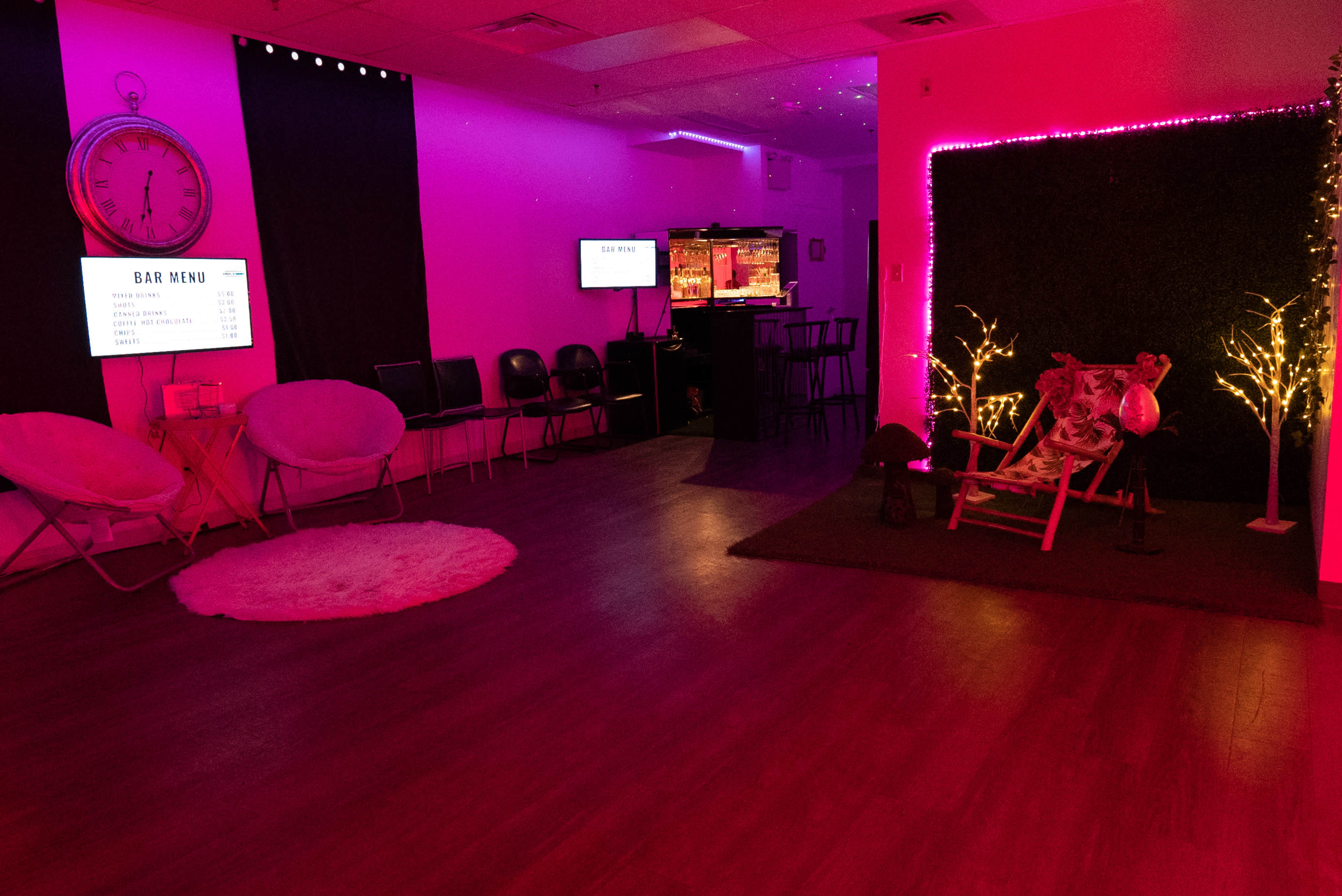 A modern interior space features pink lighting, a bar area with a menu display, and a seating arrangement with chairs and decorative elements.