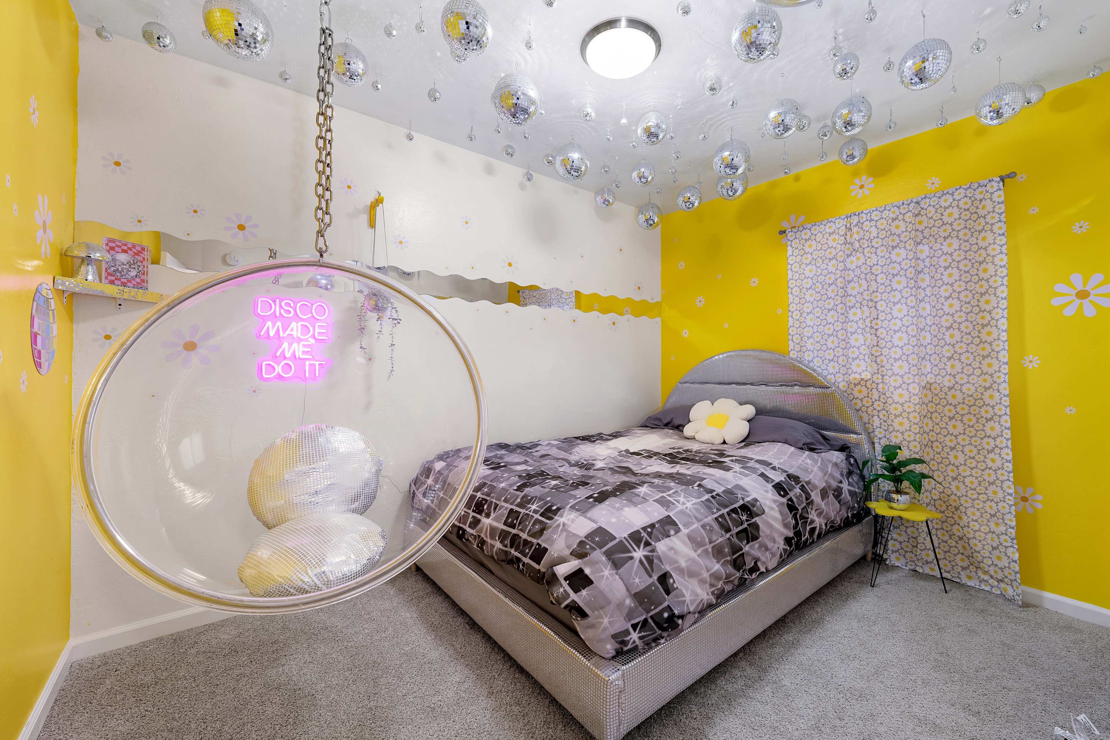 A bedroom features yellow walls adorned with flower decals, a large bed with a patterned comforter, and a hanging circular sign that reads "DISCO MADE ME DO IT," surrounded by mirrored disco balls.