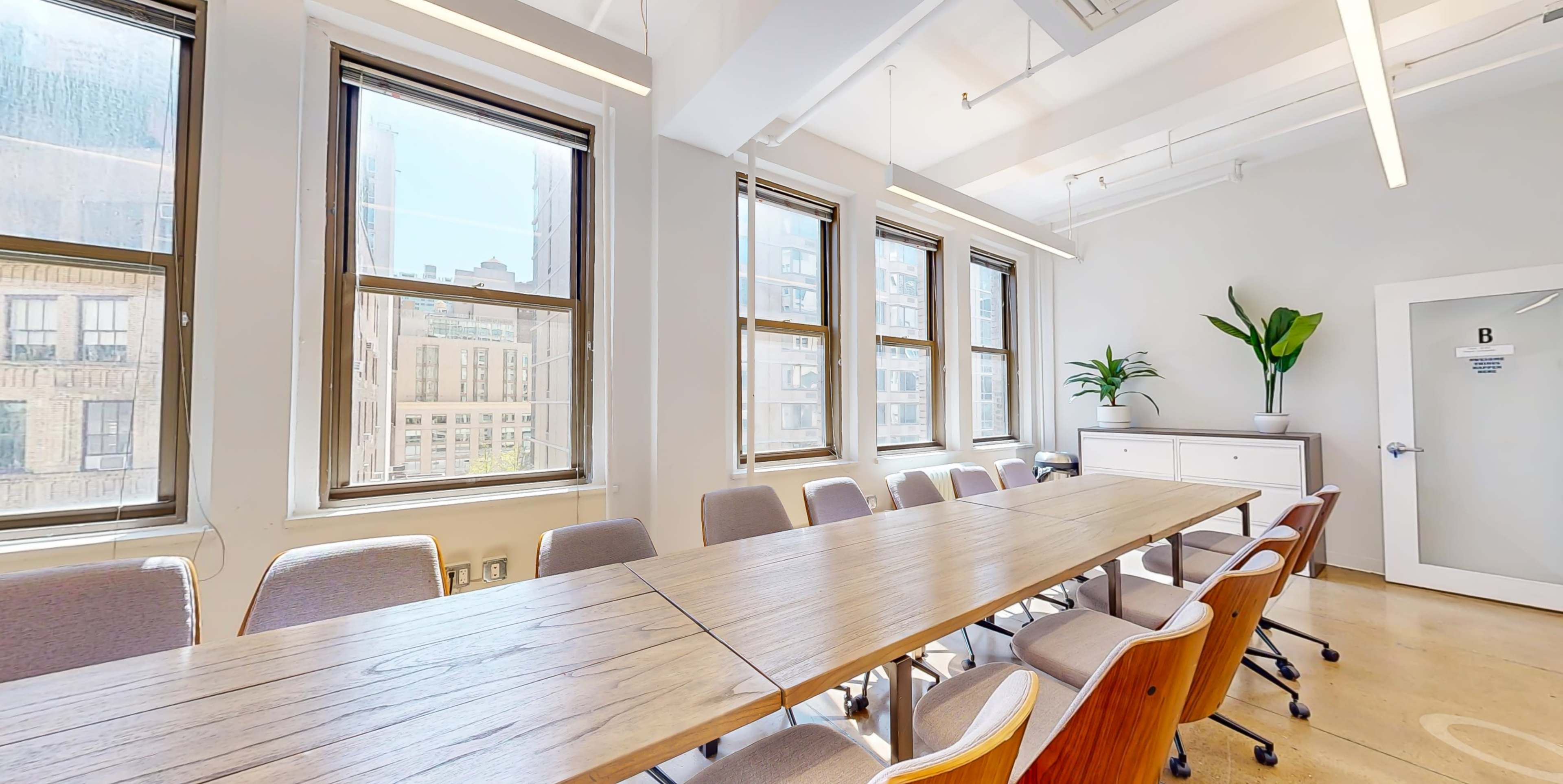 A conference room featuring a long wooden table, beige chairs, large windows, and a potted plant.