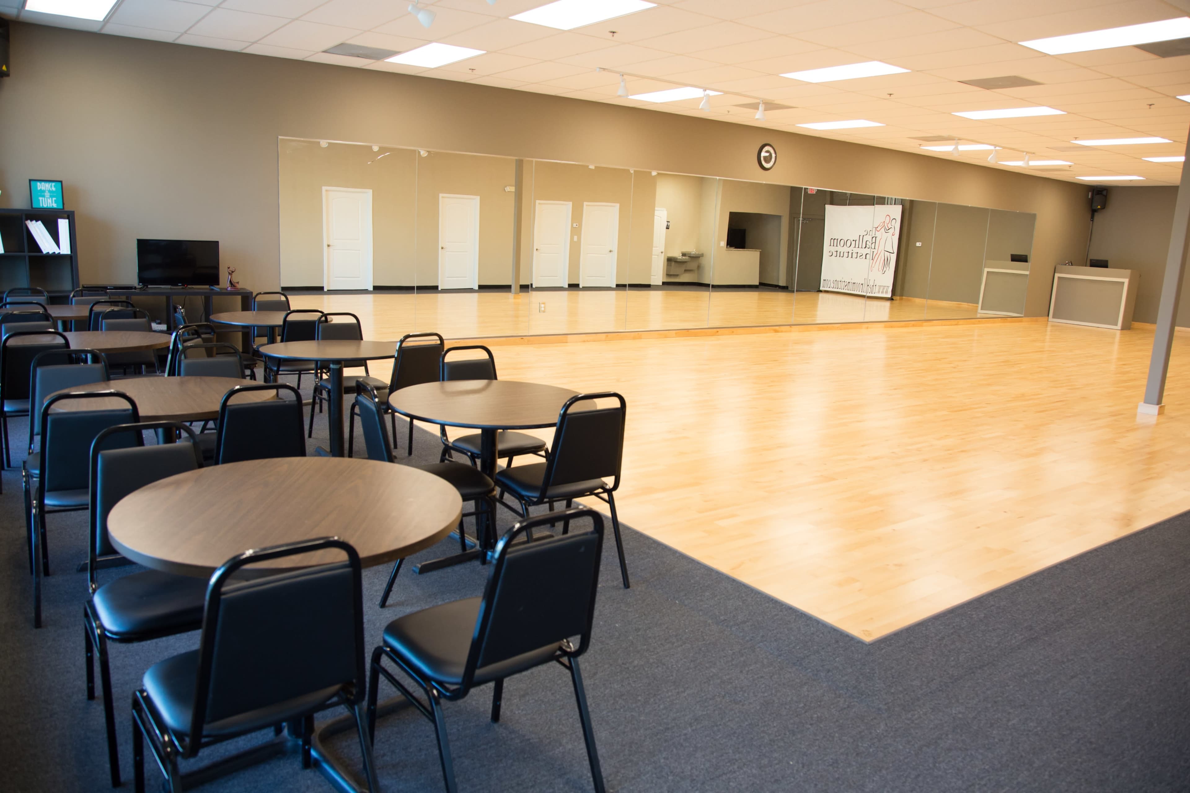 The image shows a spacious indoor studio with wooden floors, mirrored walls, several round tables with chairs, and a reception area in the background.