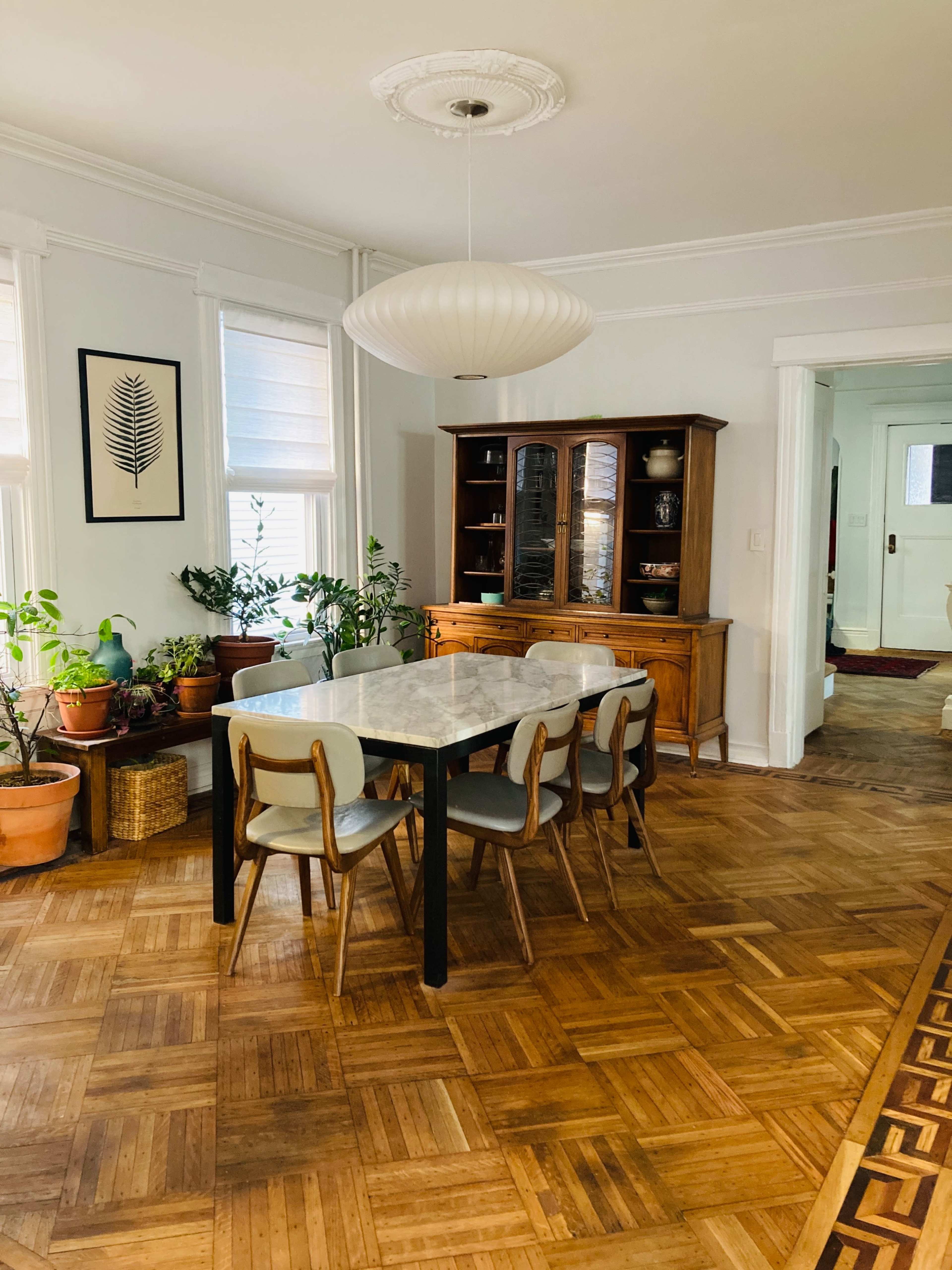 A dining room features a rectangular table with six chairs, surrounded by potted plants and a wooden cabinet against the wall.