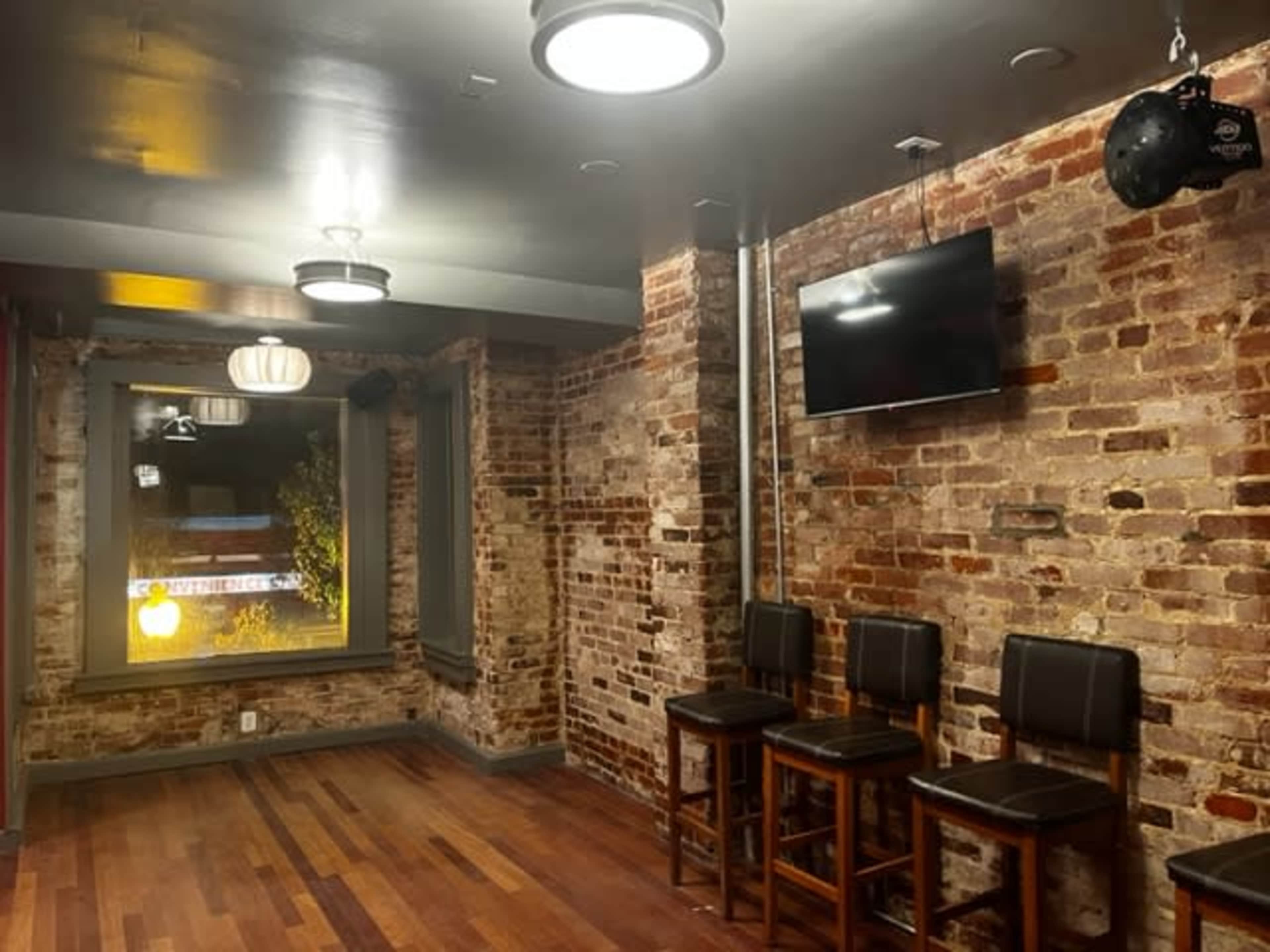 The image shows a room with exposed brick walls, wooden flooring, and a television mounted on the wall, along with a row of four wooden bar stools.