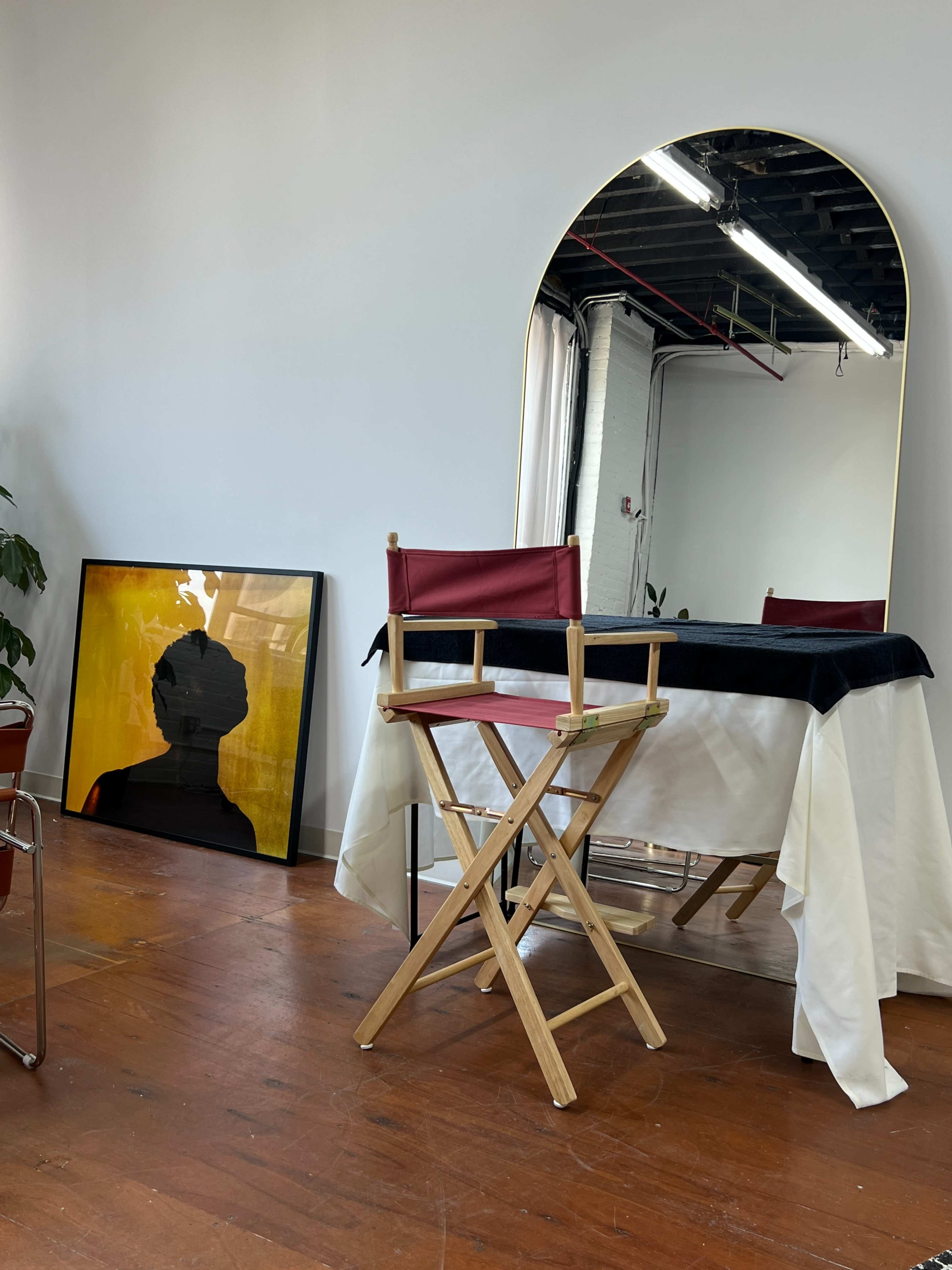 The image shows a modern, minimalistic studio space featuring a director's chair, a covered table, a large mirror, and a framed artwork on the floor.