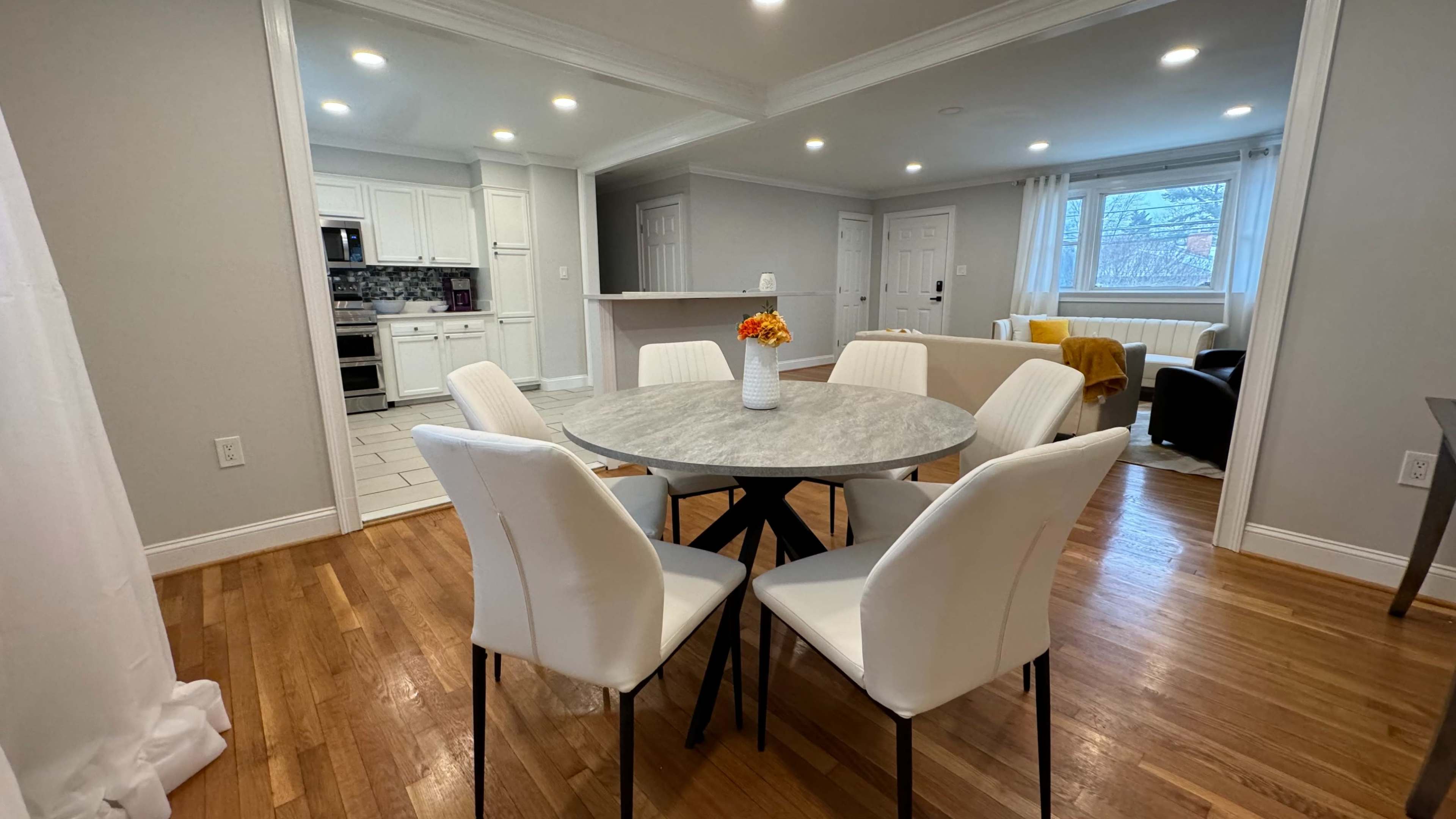 A dining area featuring a round marble table surrounded by four white chairs, with a view of a kitchen and living space in the background.