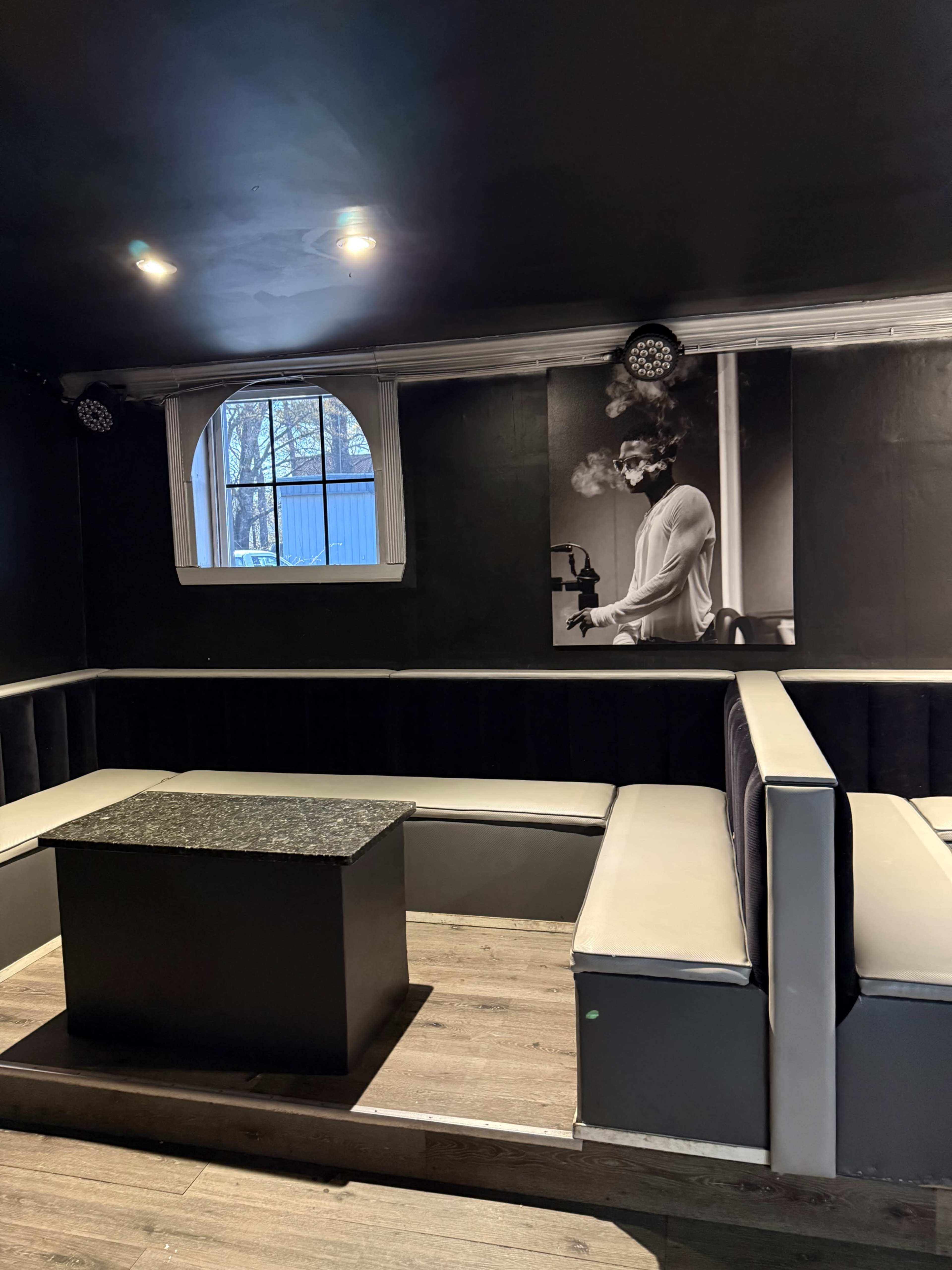 The image shows a dimly lit lounge area with black and gray furnishings, featuring a large black-and-white photograph of a person smoking on the wall.