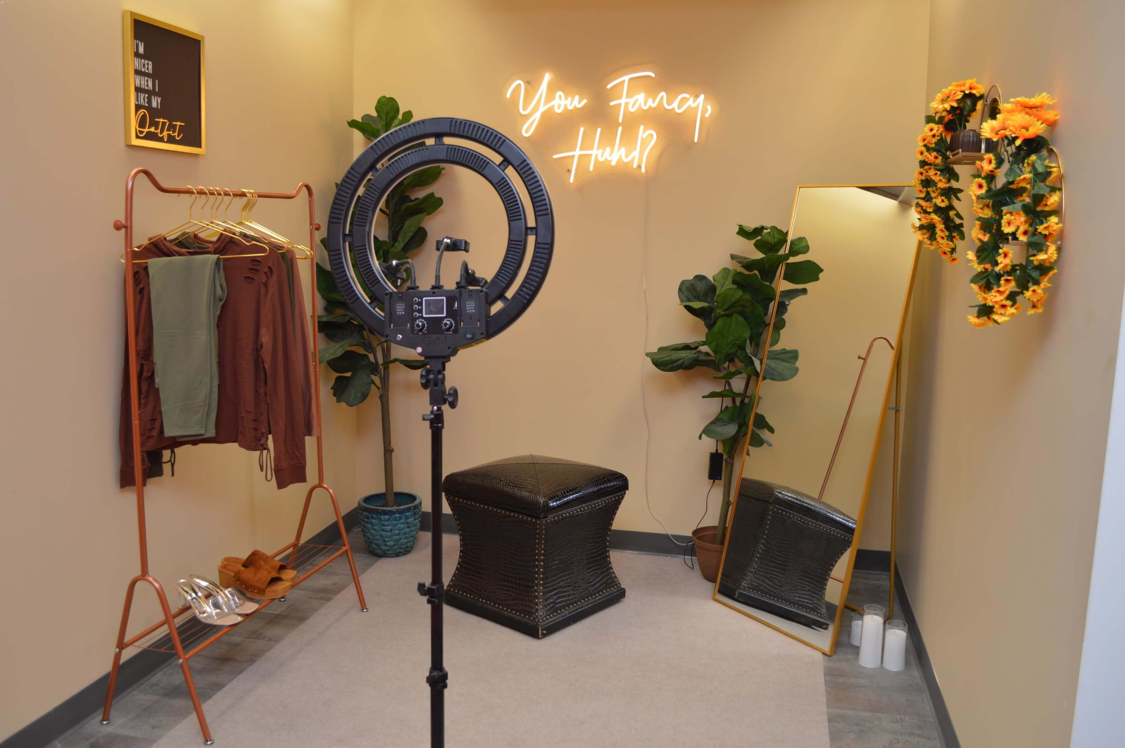 The image shows a well-decorated fitting room with a ring light, a clothing rack, a mirror, potted plants, and a neon sign that reads "You Fancy, Huh?"