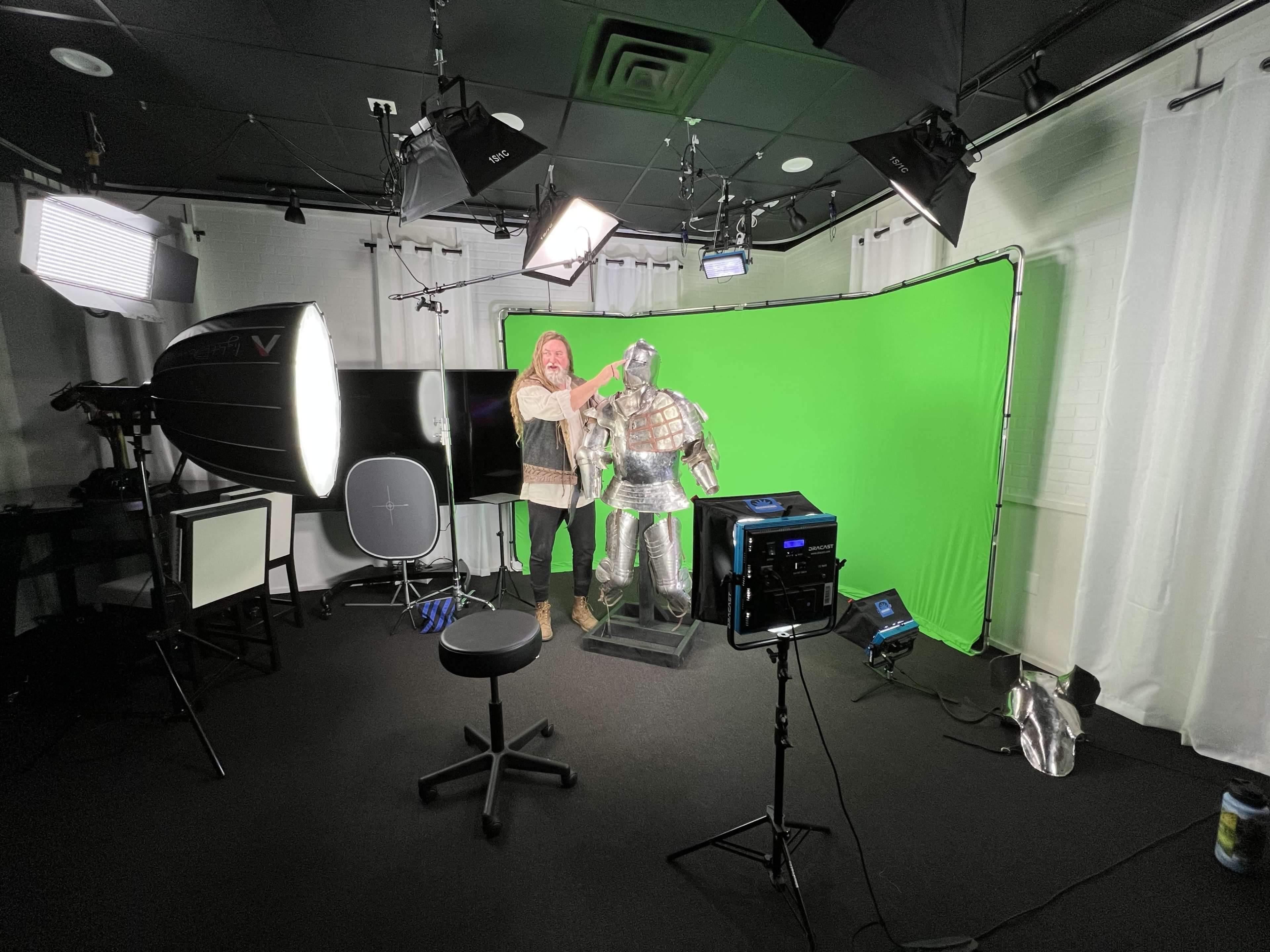 A person adjusts a suit of armor in a studio with a green backdrop and various lighting equipment.