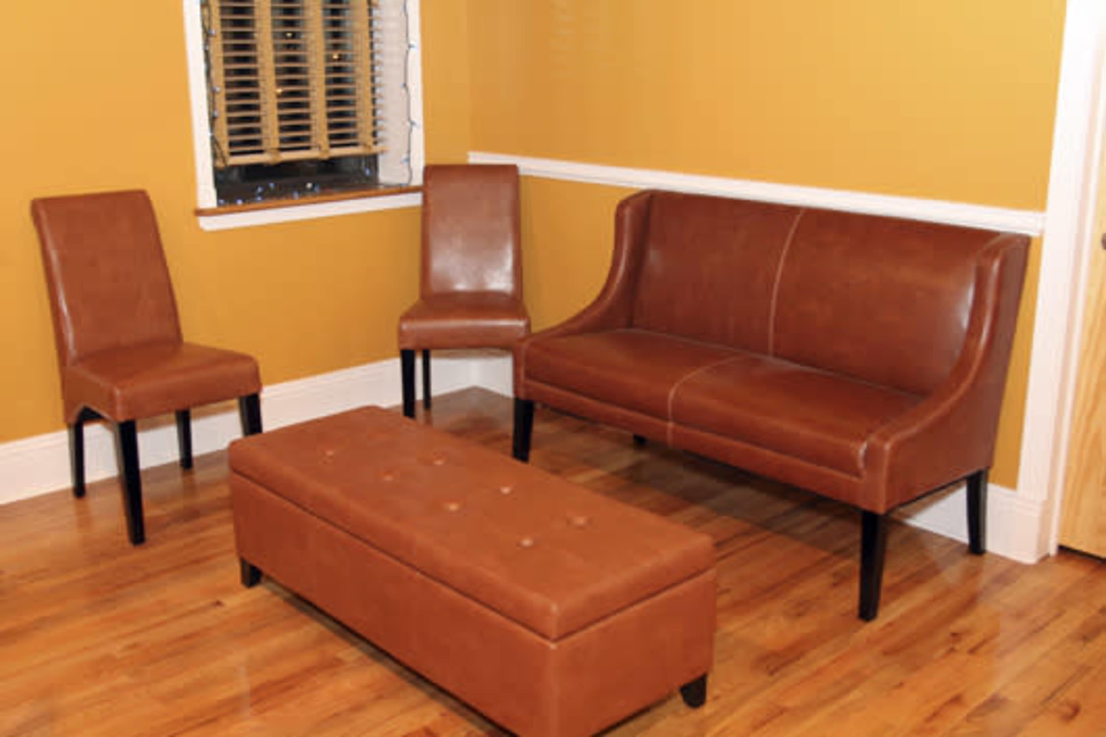 The image shows a small room with orange walls, featuring a brown leather sofa, two matching chairs, and a brown tufted bench.