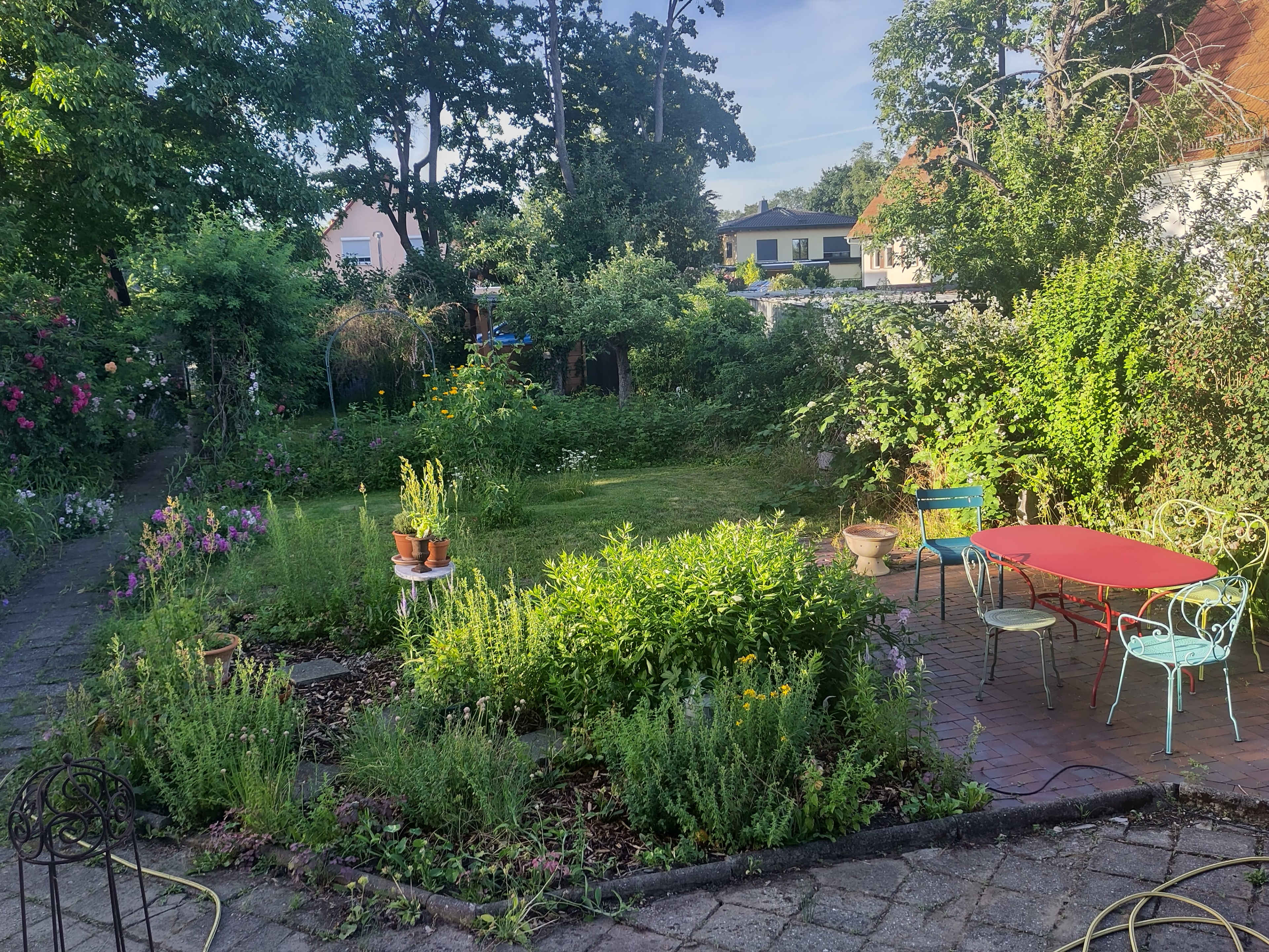 The image shows a lush garden with various plants, a grassy area, and a red table surrounded by colorful chairs.