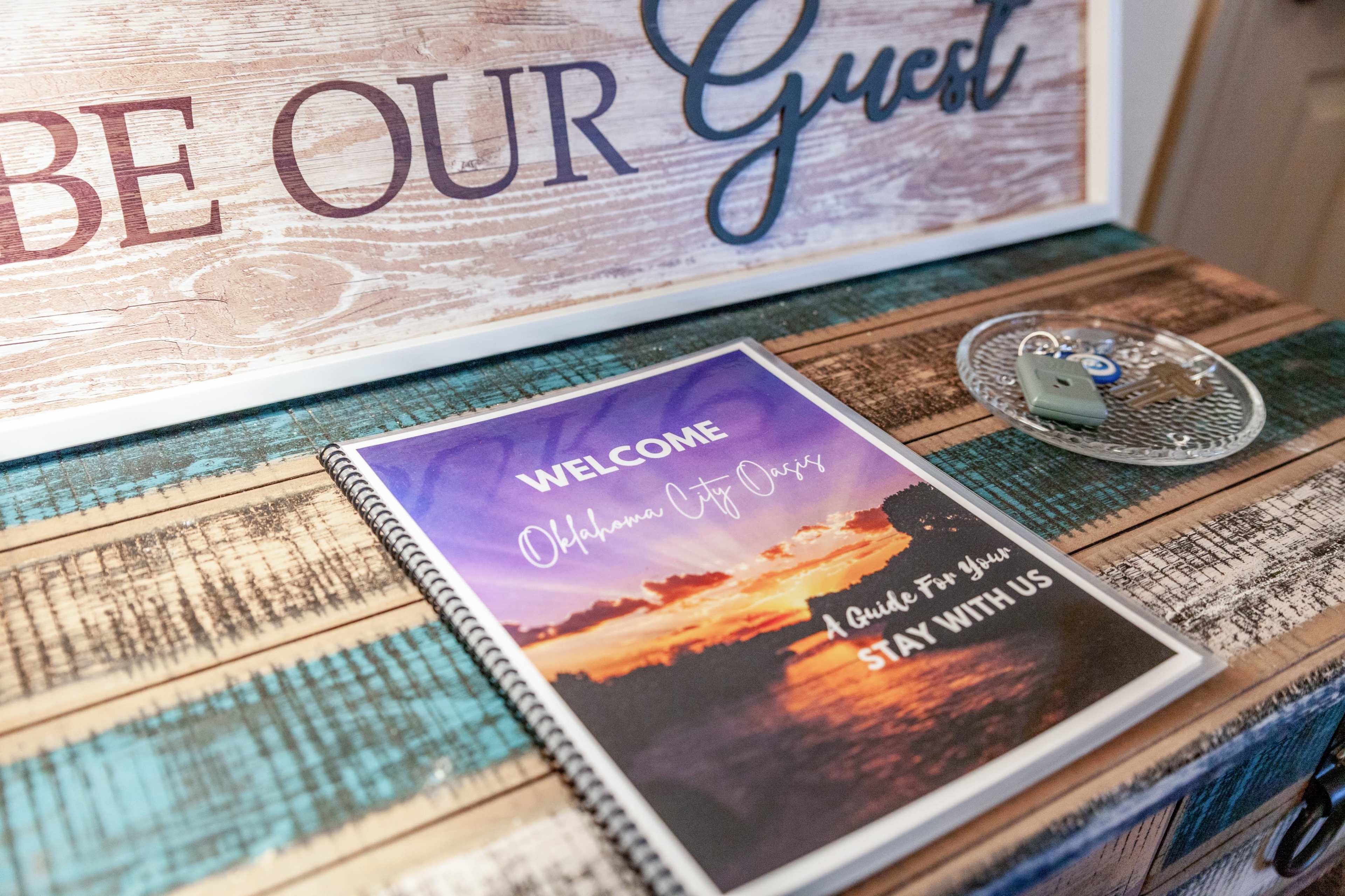 A welcome booklet titled "Oklahoma City Oasis" is placed on a wooden table, alongside a decorative key holder and a sign that says "Be Our Guest."