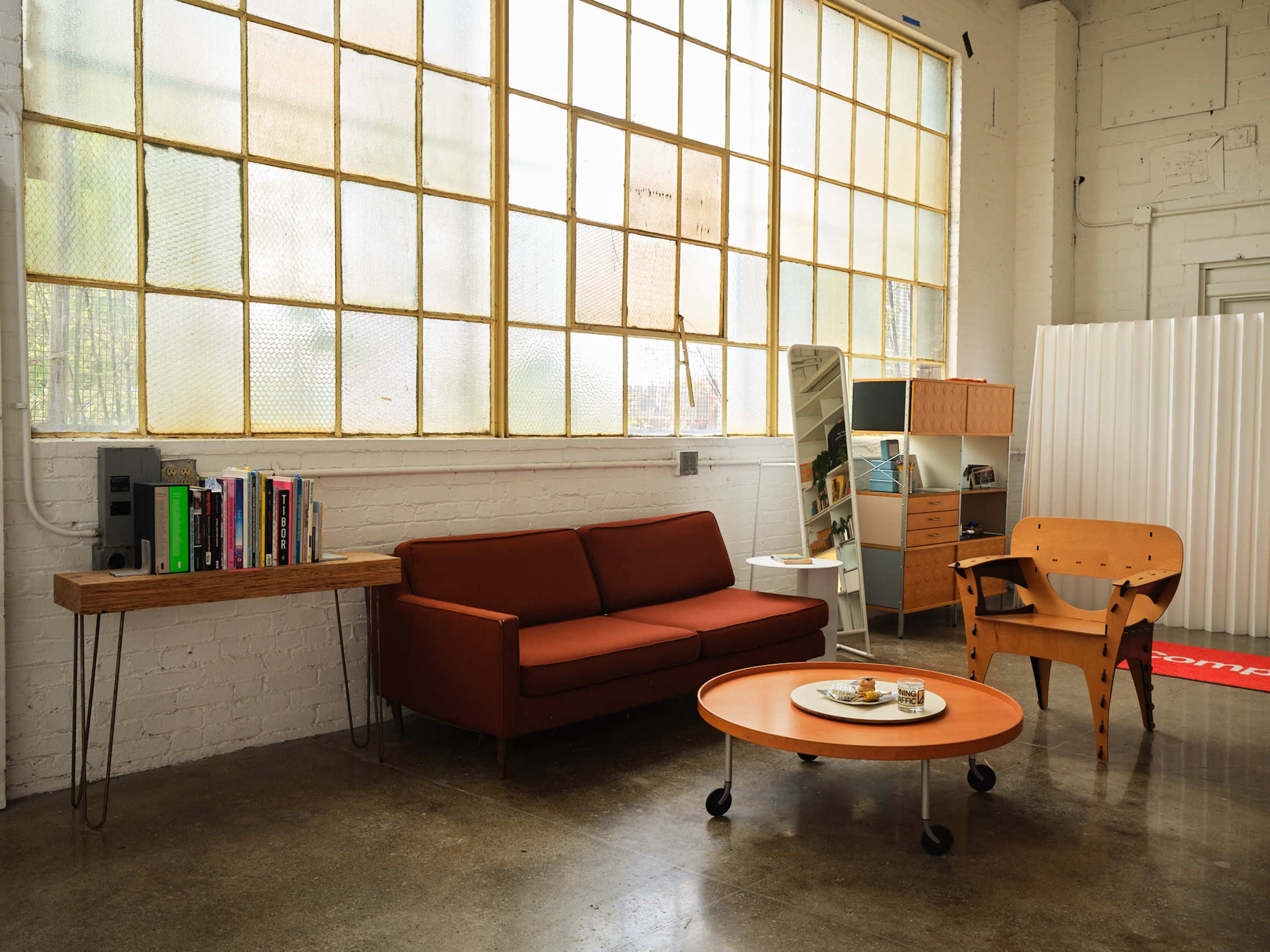 The image shows a modern living space featuring a brown sofa, a circular coffee table with snacks, and a wooden bookshelf beside large windows.