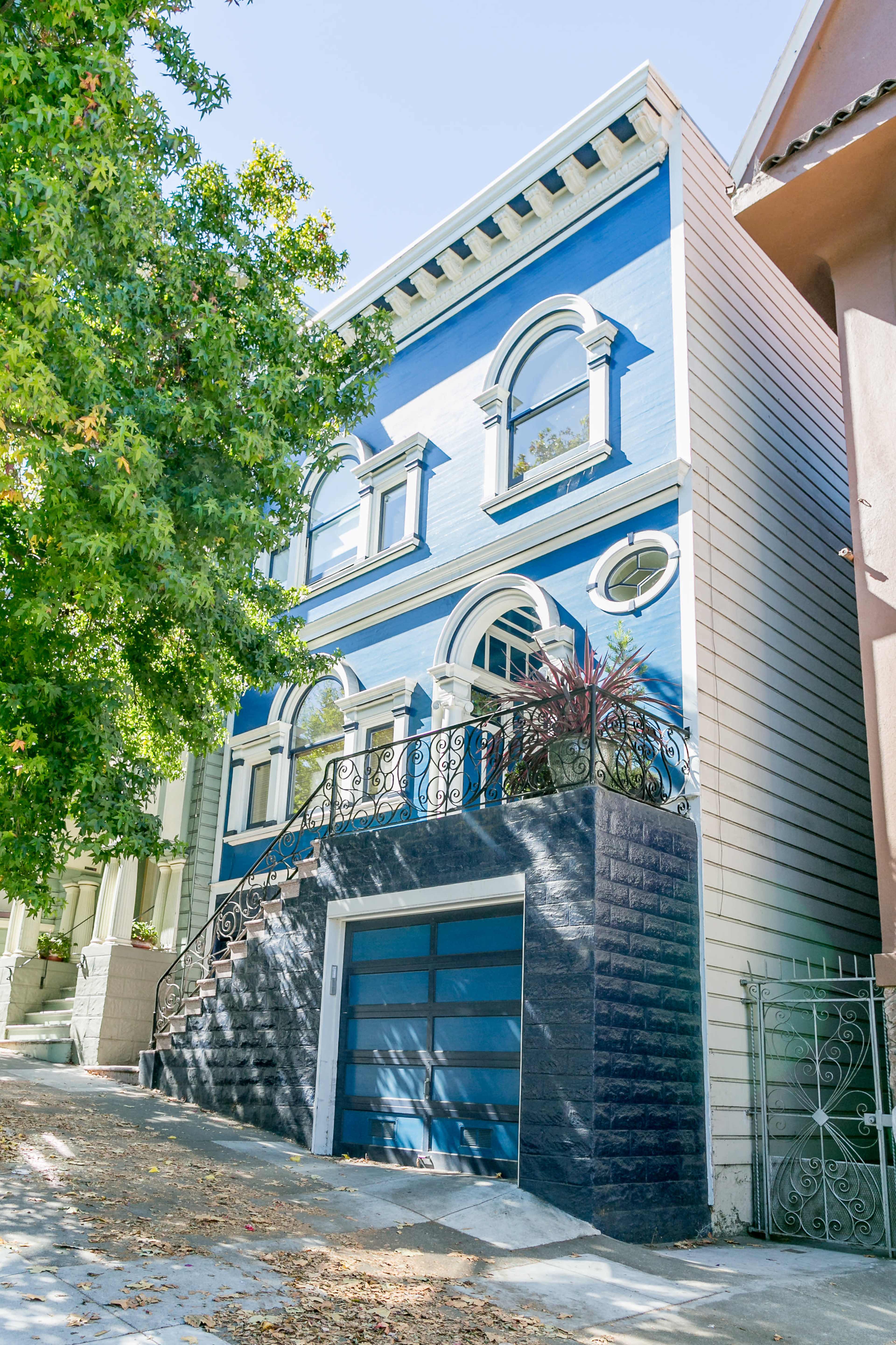 A three-story blue house with decorative windows and a dark stone garage door is situated on a sloped driveway, surrounded by greenery.