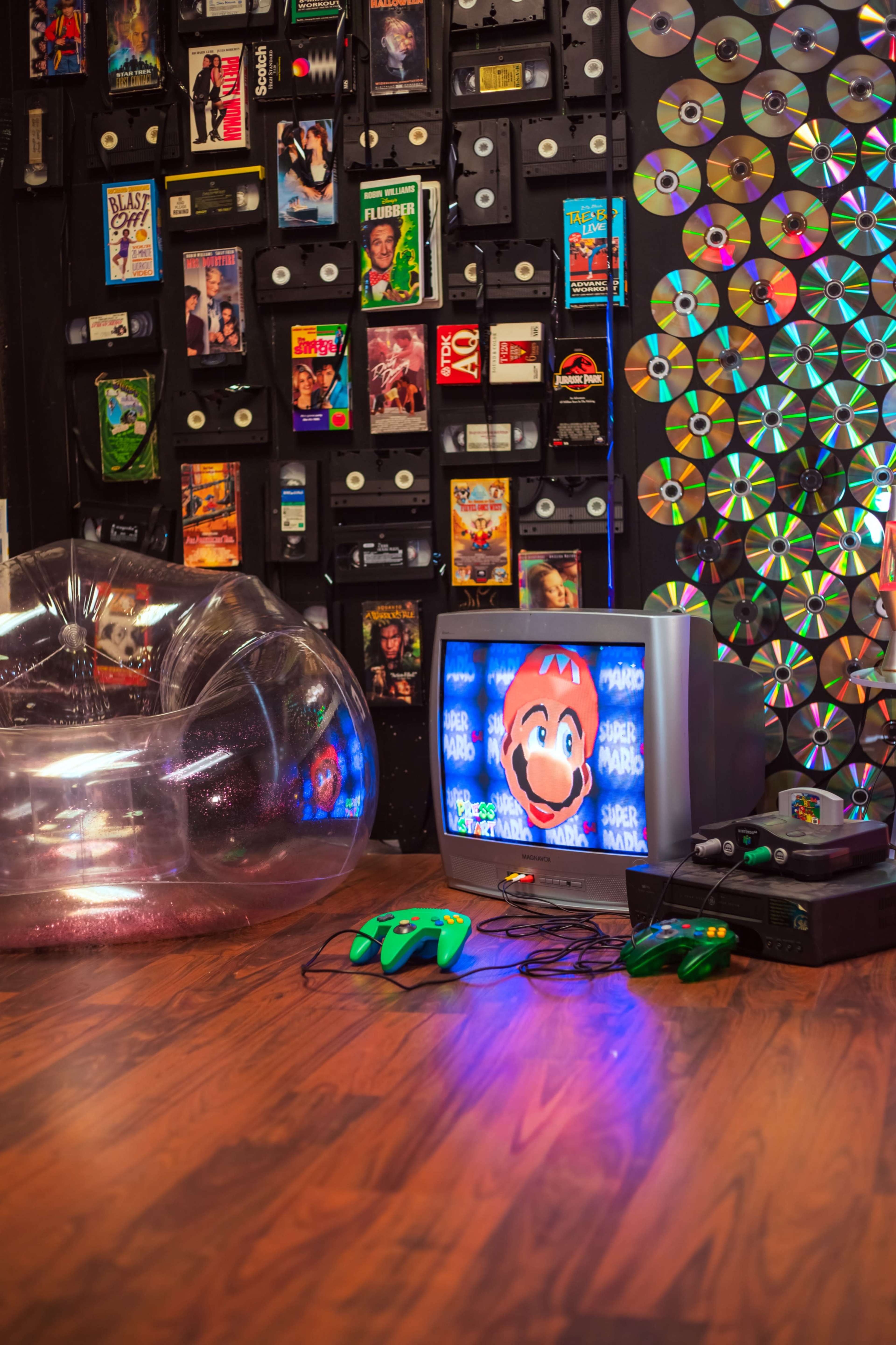 The scene features a retro video gaming setup with a television displaying "Super Mario" and various gaming consoles and memorabilia against a wall of VHS tapes and CDs.