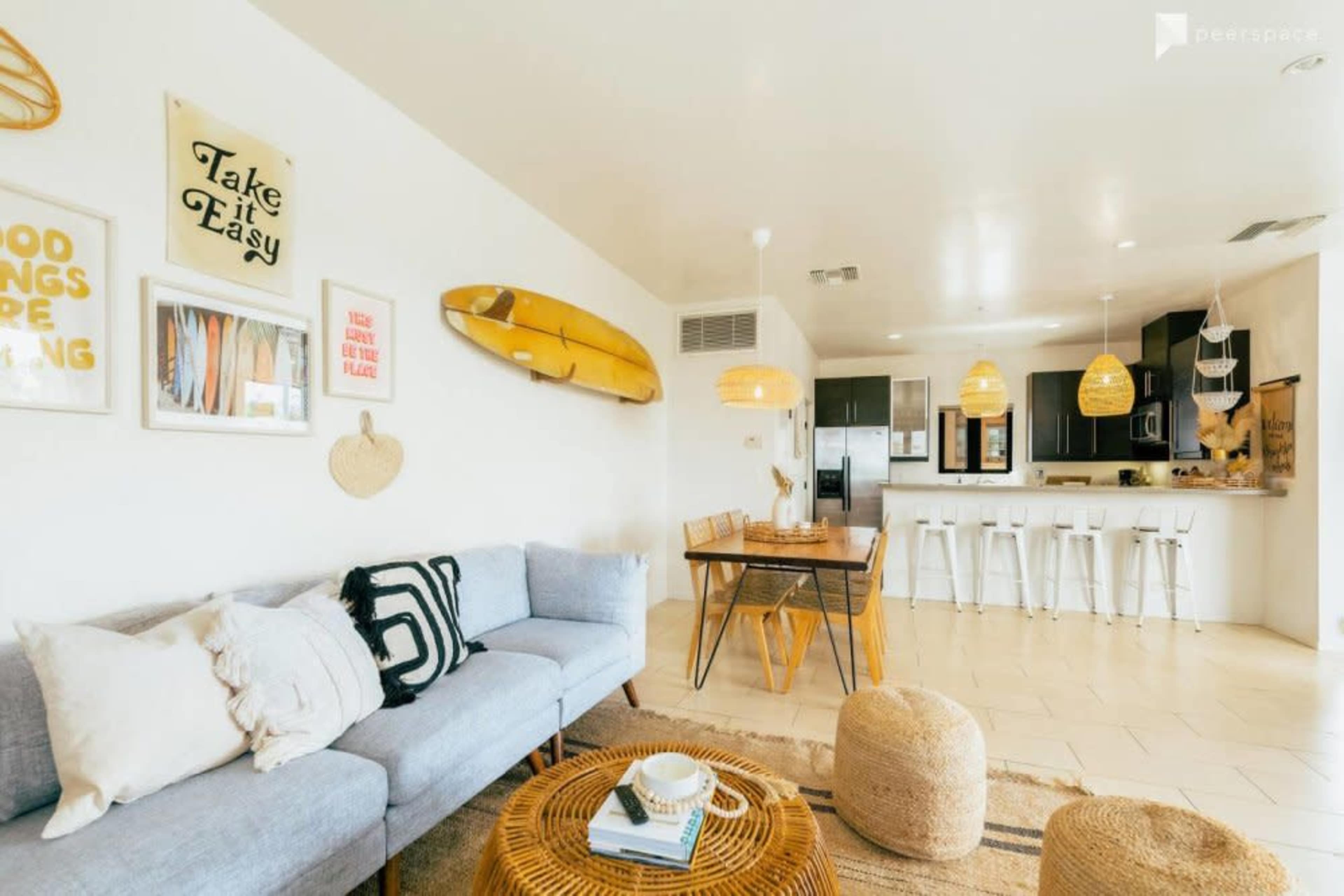The image shows a bright, open-concept living area with a blue couch, a wooden dining table, and a kitchen in the background featuring modern appliances and a surfboard decoration on the wall.