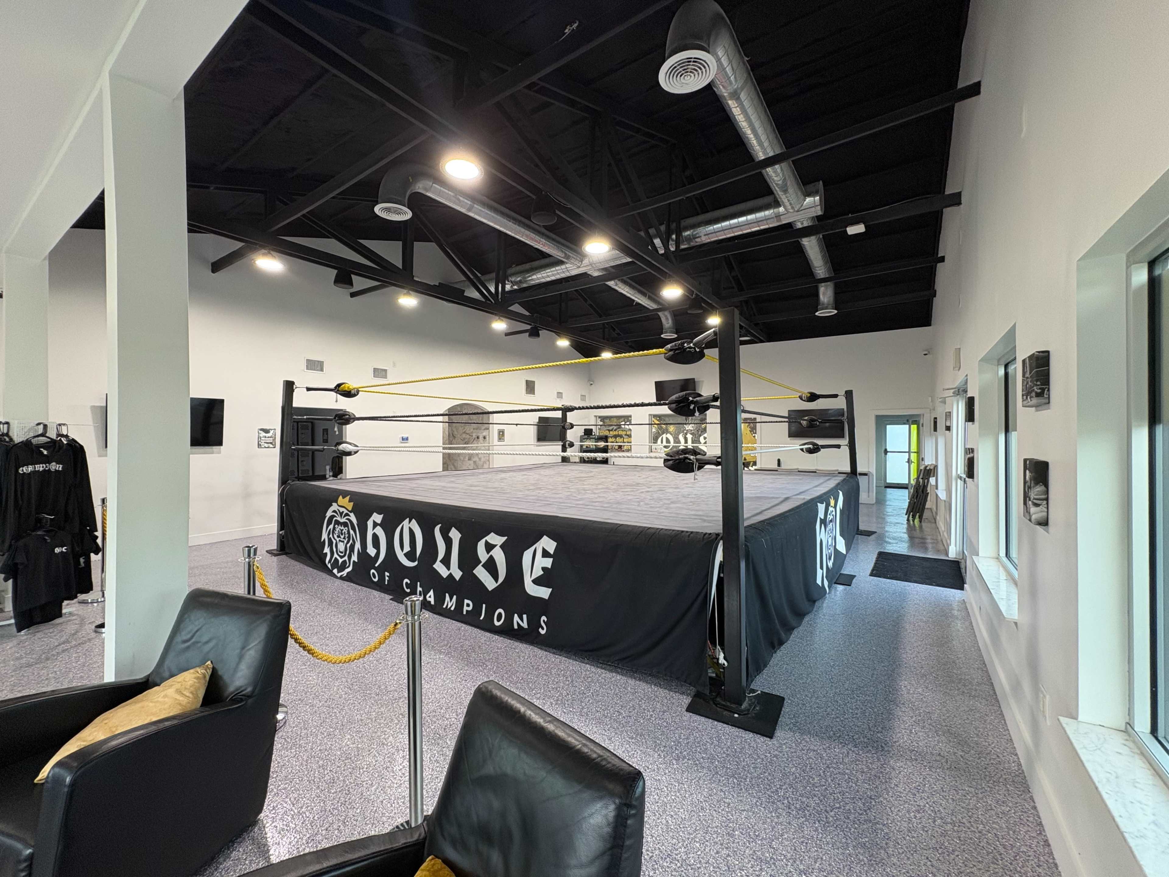 The image shows a professional wrestling training facility featuring a wrestling ring with yellow ropes, surrounded by sleek furnishings and bright white walls.
