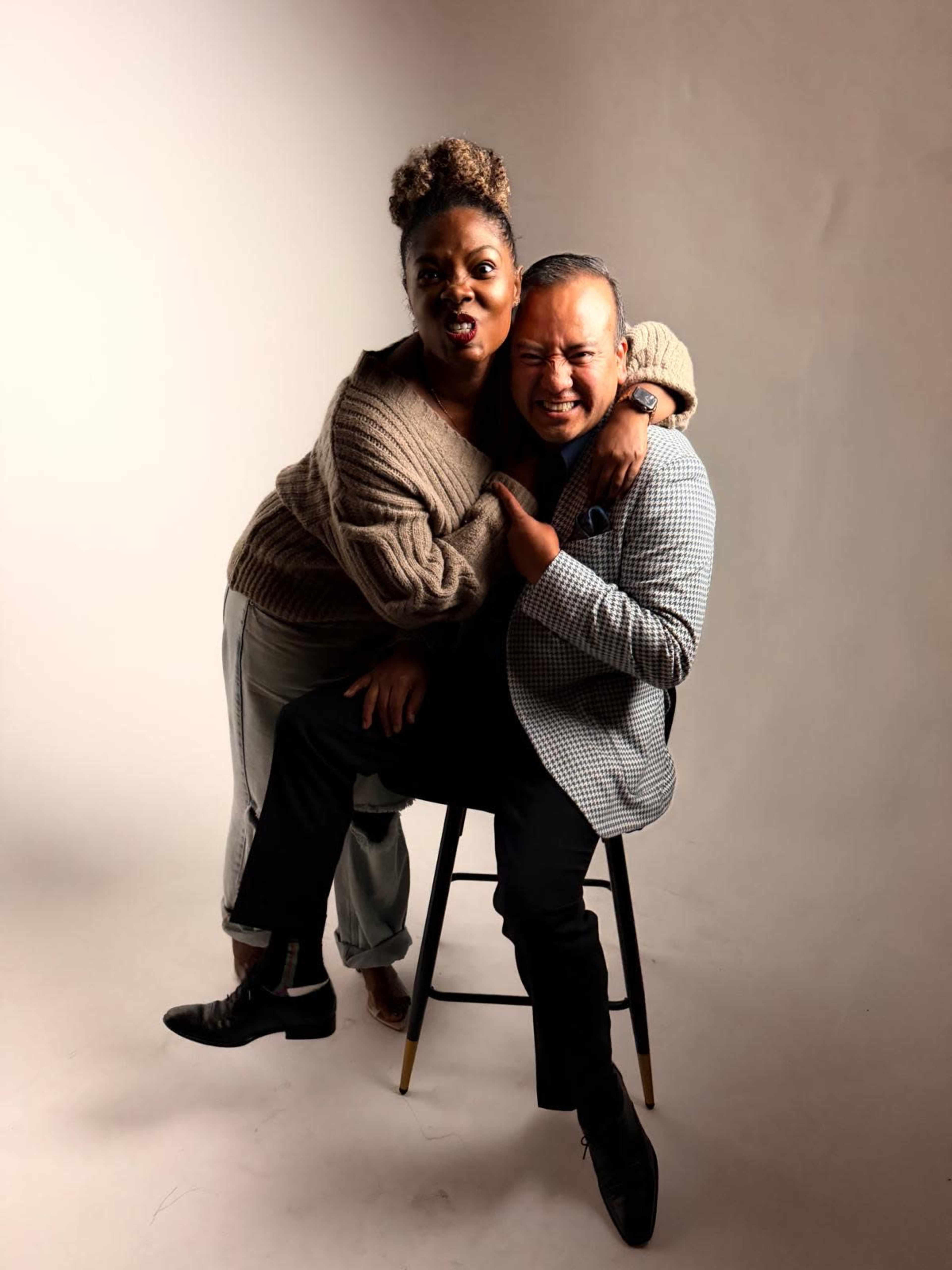 A woman playfully sits on the lap of a man, both smiling while posing for the camera in a studio setting.