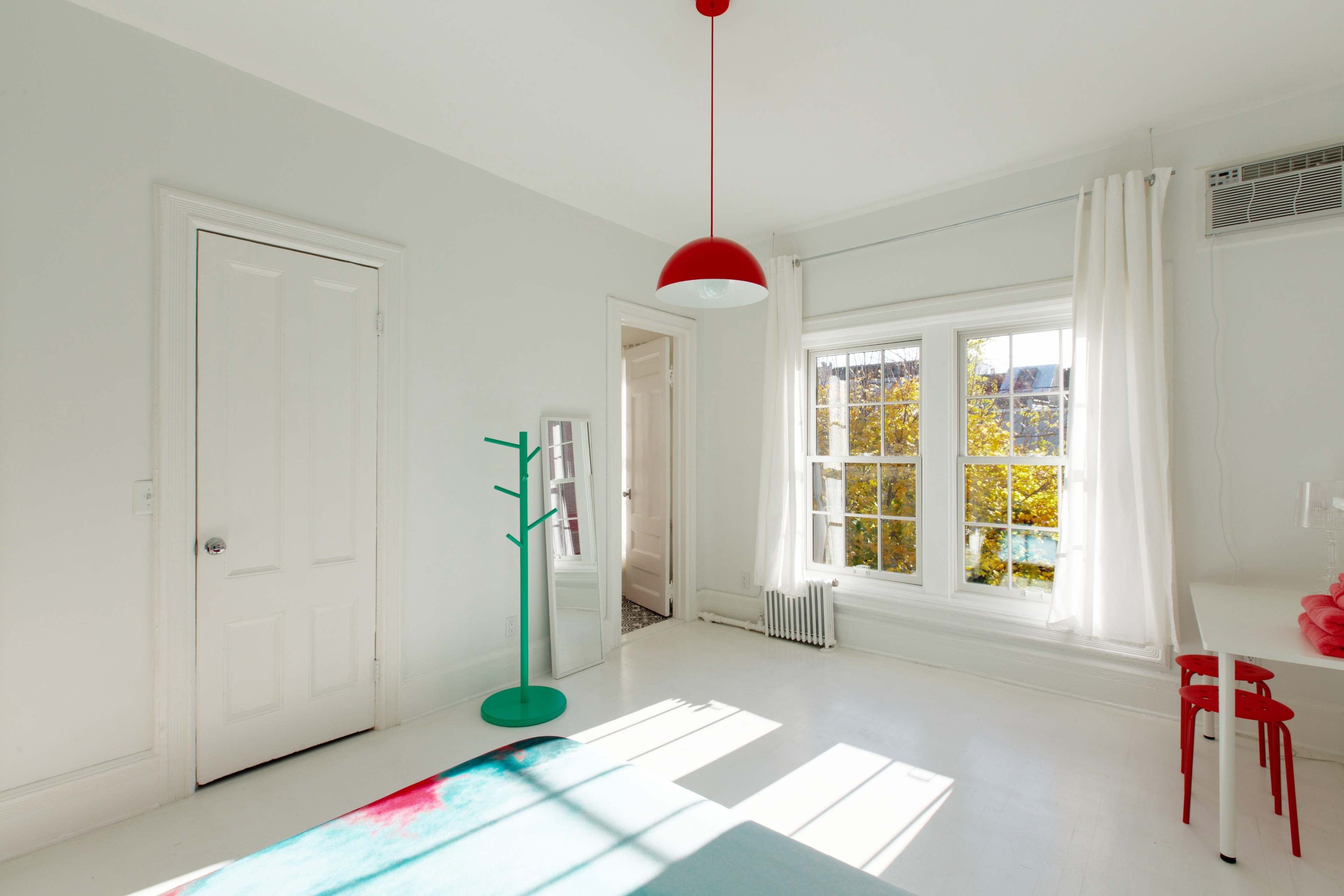 A bright, minimalistic bedroom with a white color scheme, featuring a red pendant light, a green coat rack, and large windows that let in natural light.