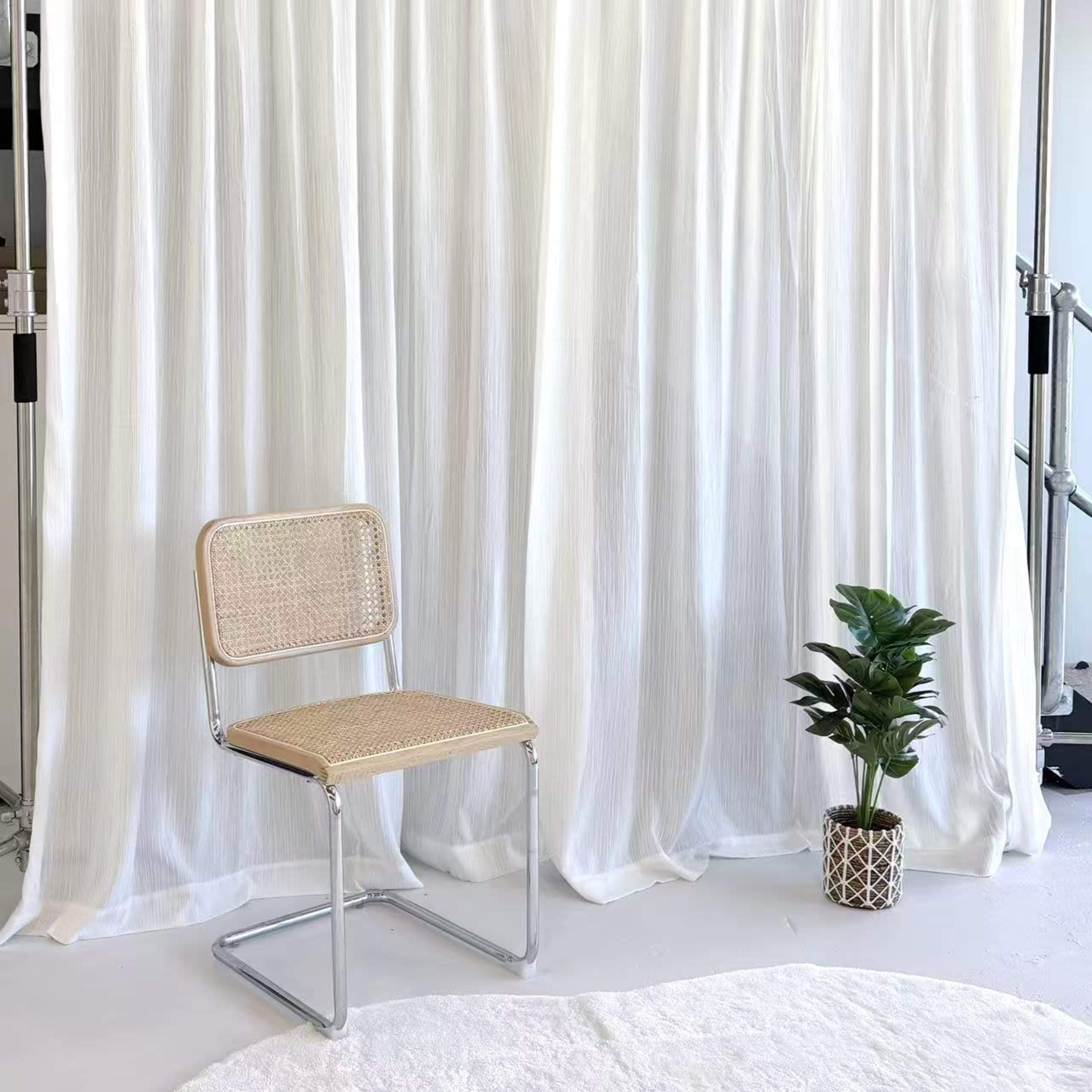 The image features a simple chair with a wicker seat in front of a white curtain, alongside a small potted plant on the floor.