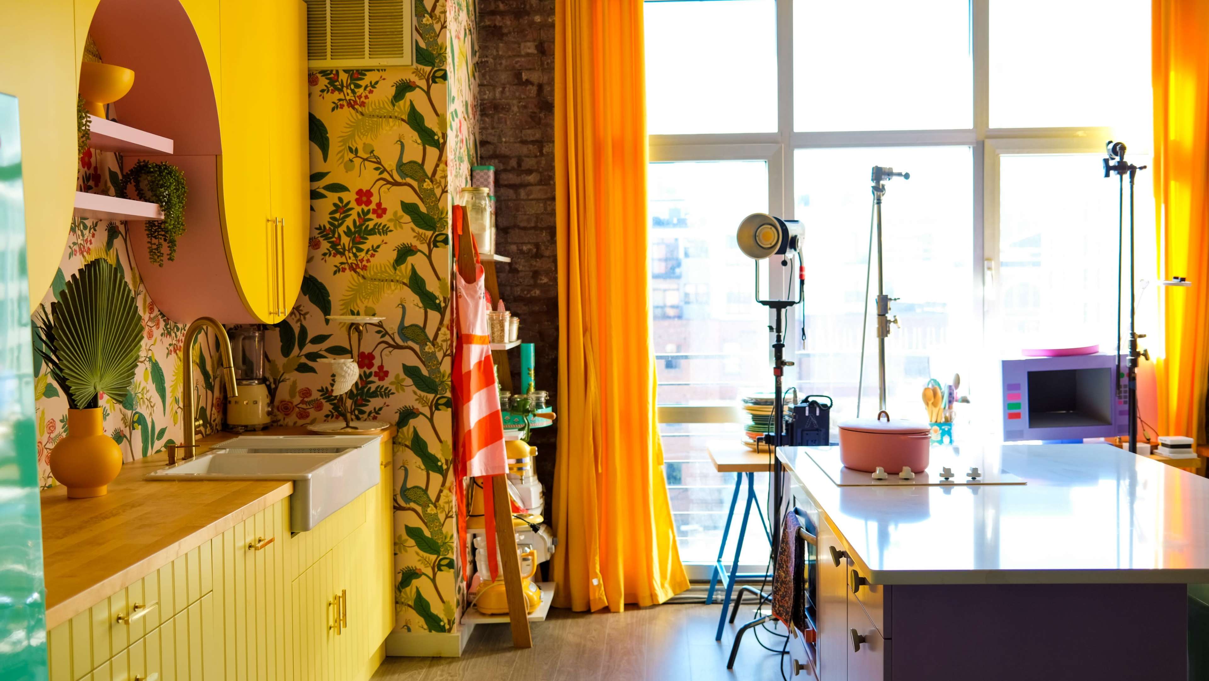 A brightly colored kitchen features yellow cabinets, a floral wallpaper, and large windows with orange curtains.