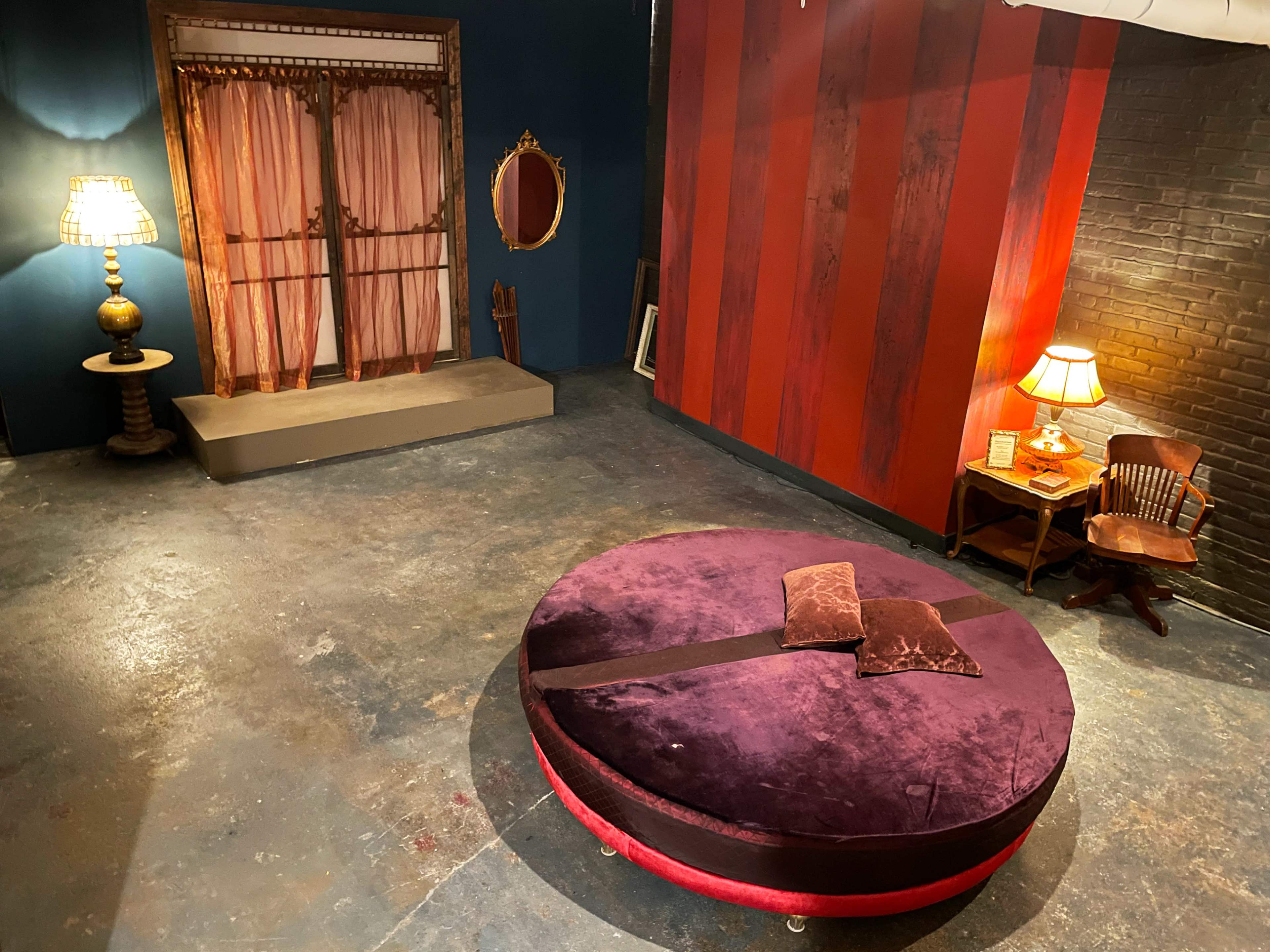 The image shows a room with a round purple seating area in the center, a red-striped wall, a vintage lamp, and a small table with a chair in one corner.