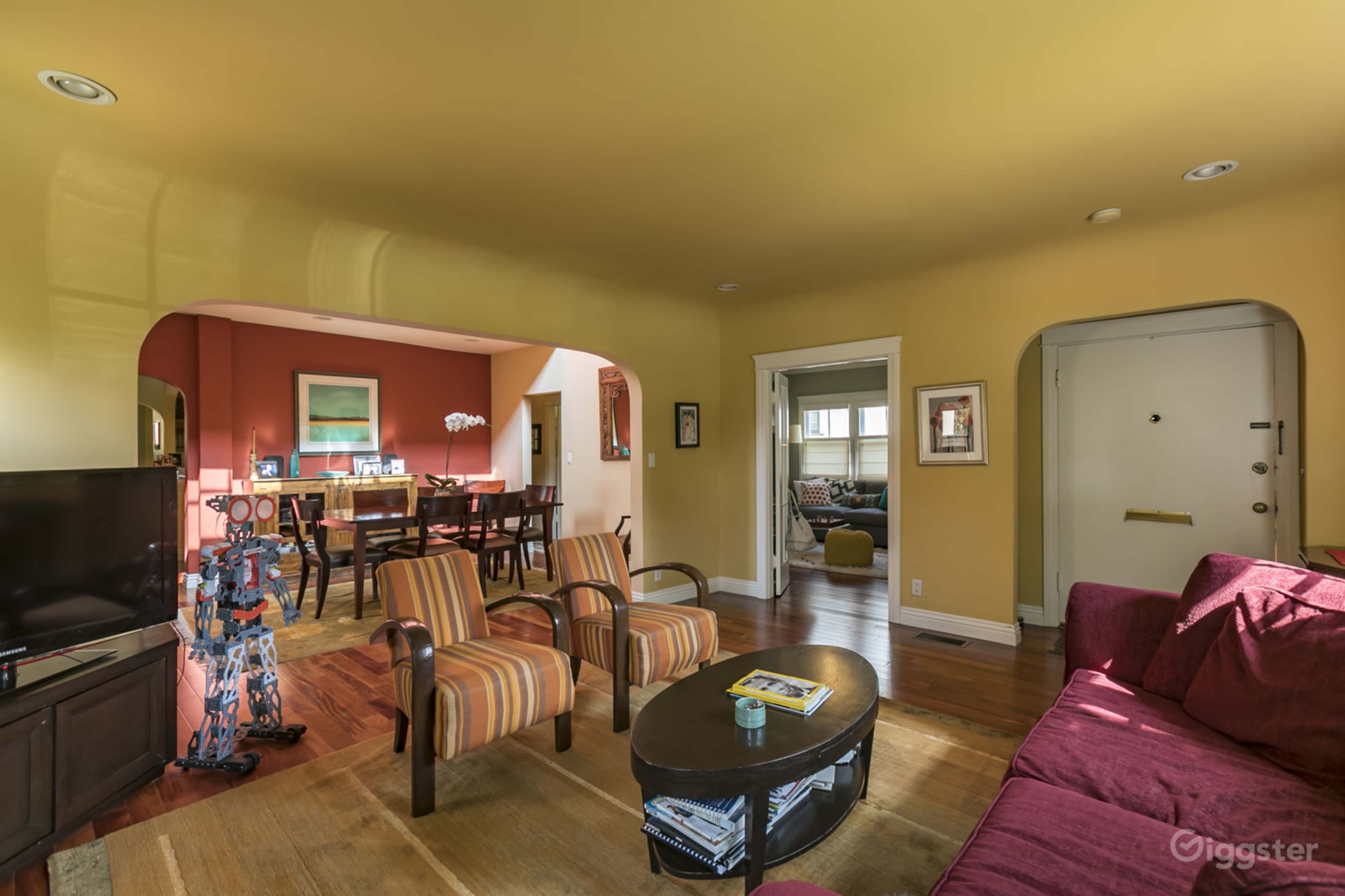 The image shows a living room with yellow walls, a red accent wall, a brown sofa, striped armchairs, and a dining area visible in the background.