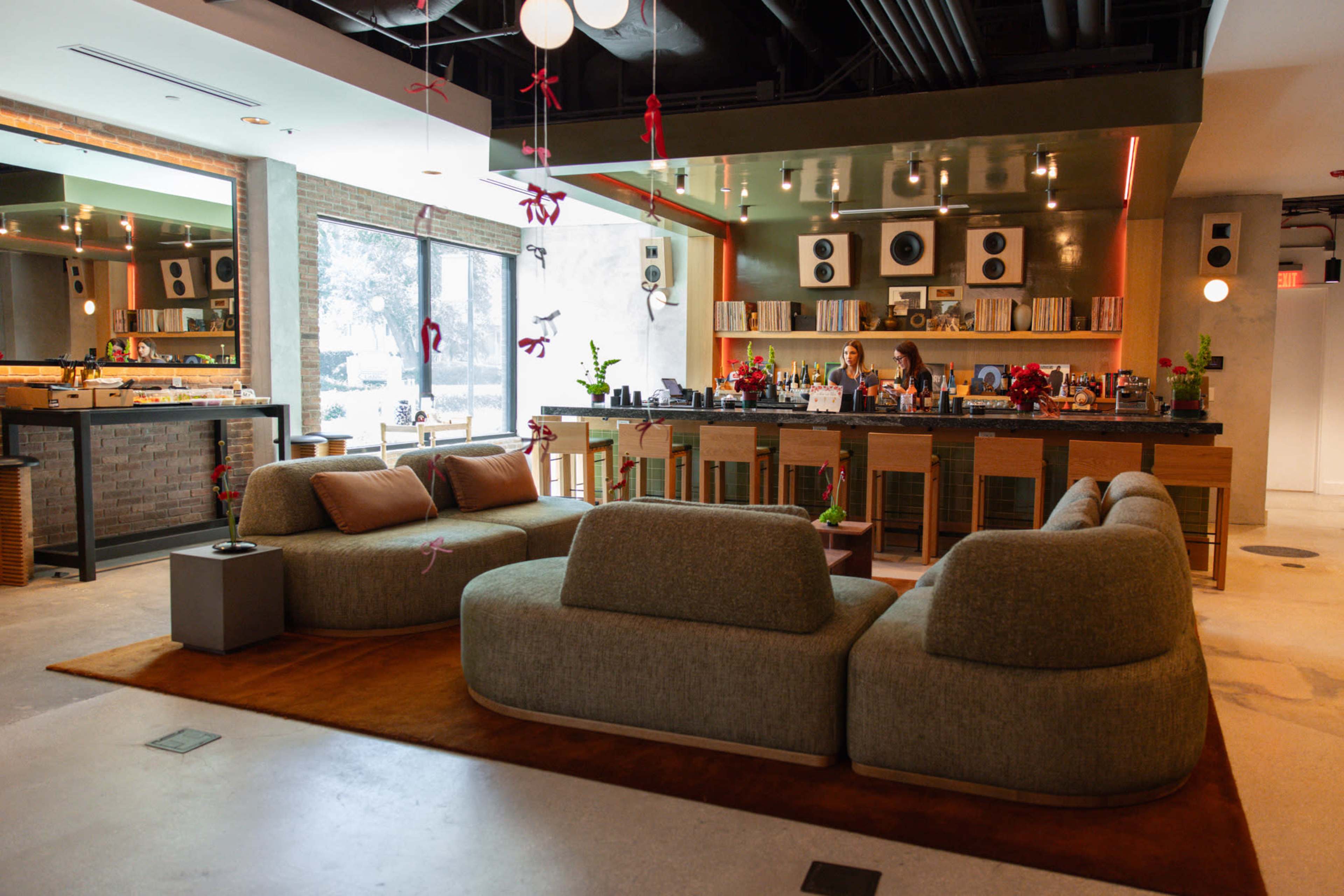 The image shows a modern lounge area featuring a curved sofa, a bar with speakers behind it, and large windows letting in natural light.